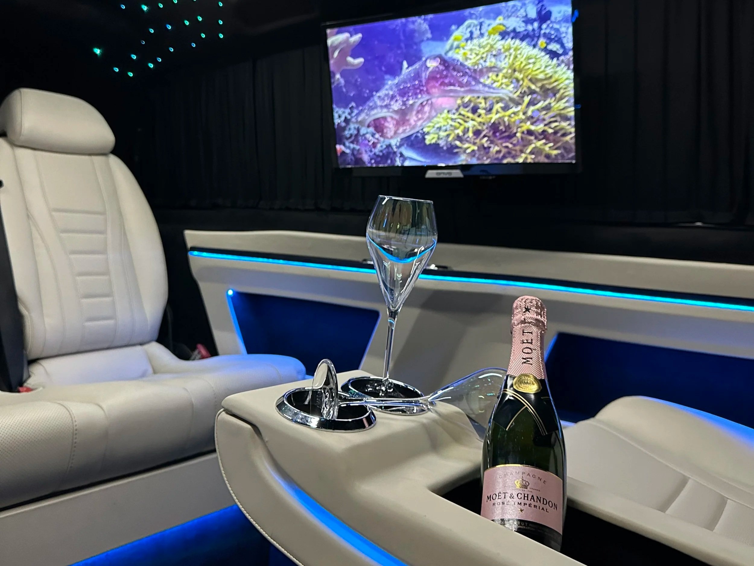 Luxury car interior with white leather seating, a bottle of Moët & Chandon champagne, two champagne glasses, and a TV screen displaying an underwater scene with a fish and coral.
