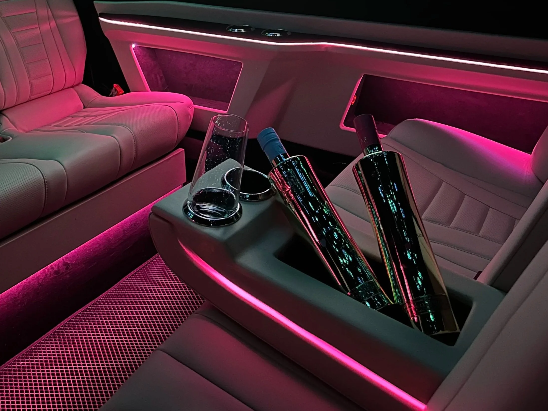 Luxury car interior with pink neon lighting, white leather seats, a table with a wine glass and three bottles, and a pink-lit geometric floor mat.