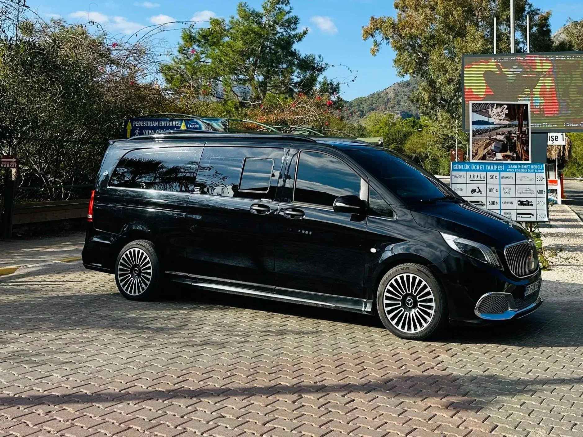 Black luxury van parked in a lot with trees, hills, and billboards in the background.
