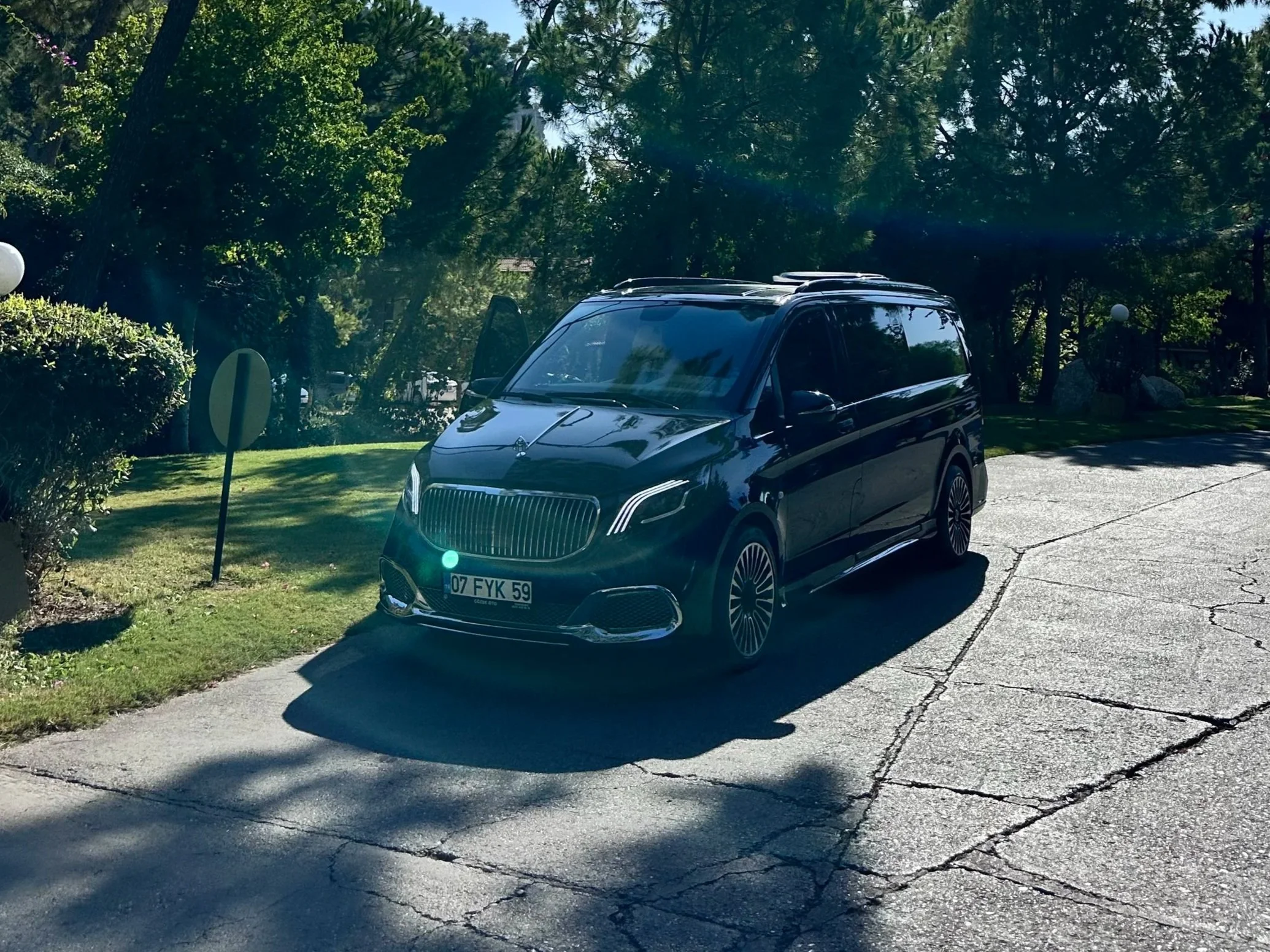 A black Mercedes-Benz van parked on a cracked driveway in a lush, green yard with trees and bushes, under a bright sunny sky.