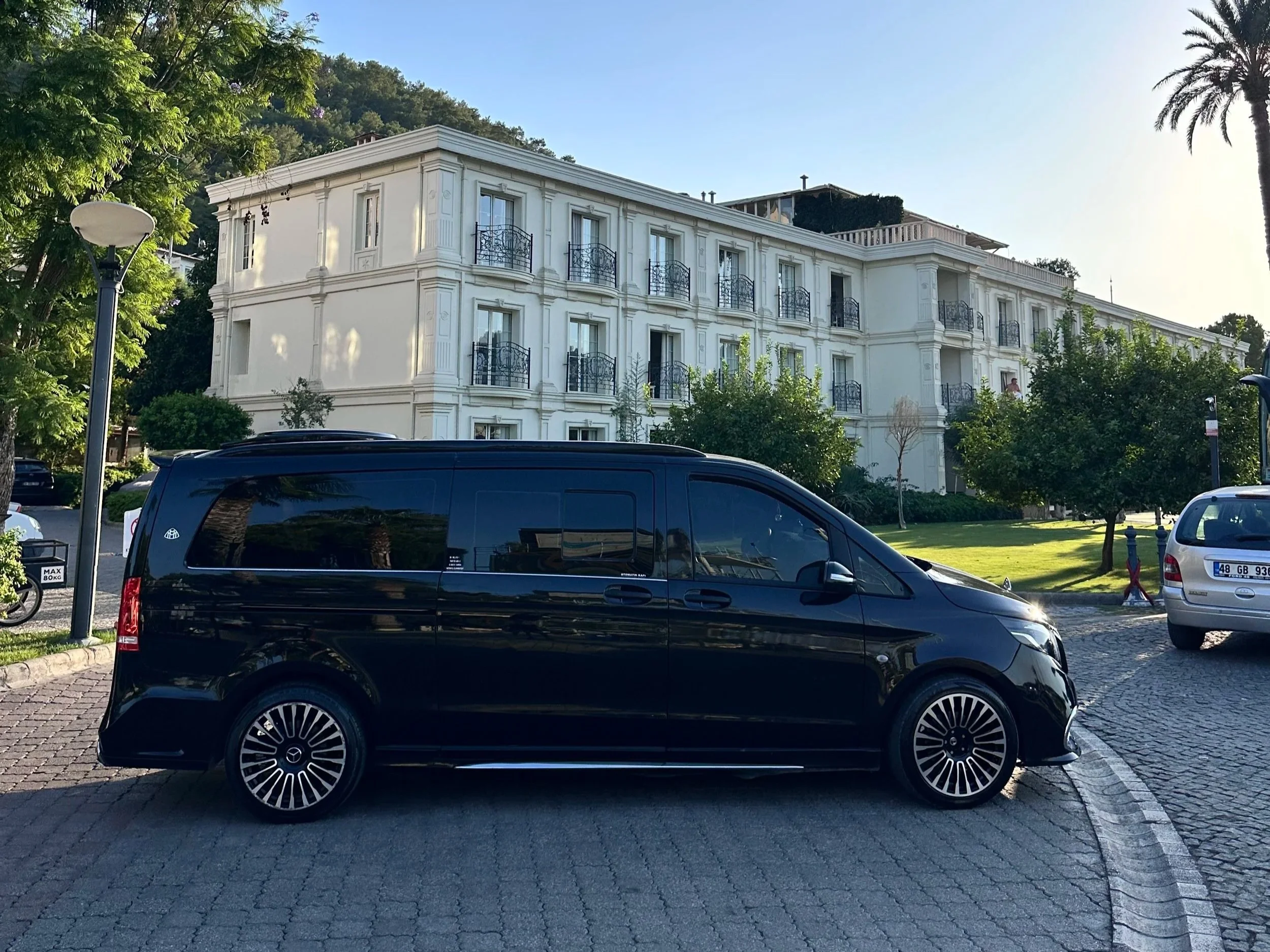 Black van parked on a cobblestone street in front of a white multi-story building with balconies, surrounded by green trees and a grassy area, in sunny weather.