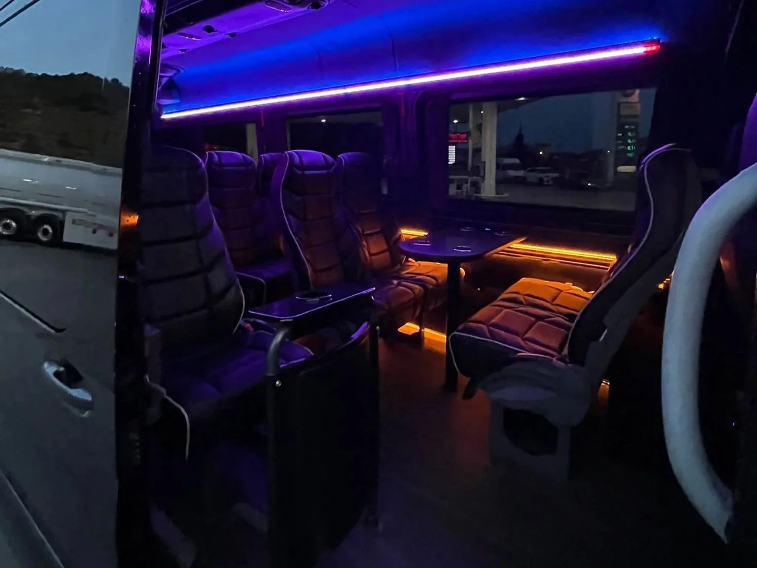 Inside a bus with black leather seats illuminated by purple and orange LED lights, with a window showing an outdoor parking lot at dusk.