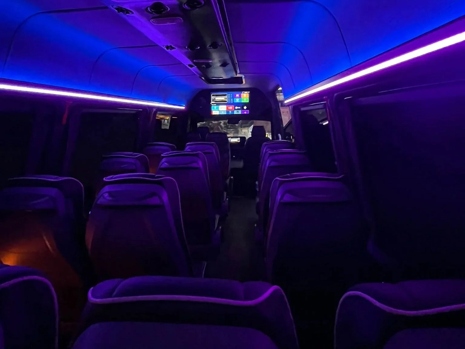 Empty bus interior illuminated with purple and blue LED lighting, showing multiple seats facing forward, and a digital screen at the front displaying various icons and information.