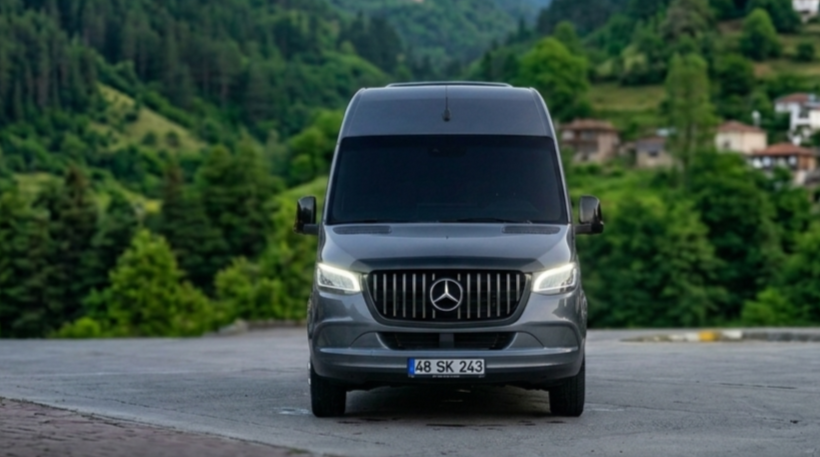 A black Mercedes-Benz van parked on a paved area in front of a green, hilly landscape with trees and houses in the background.