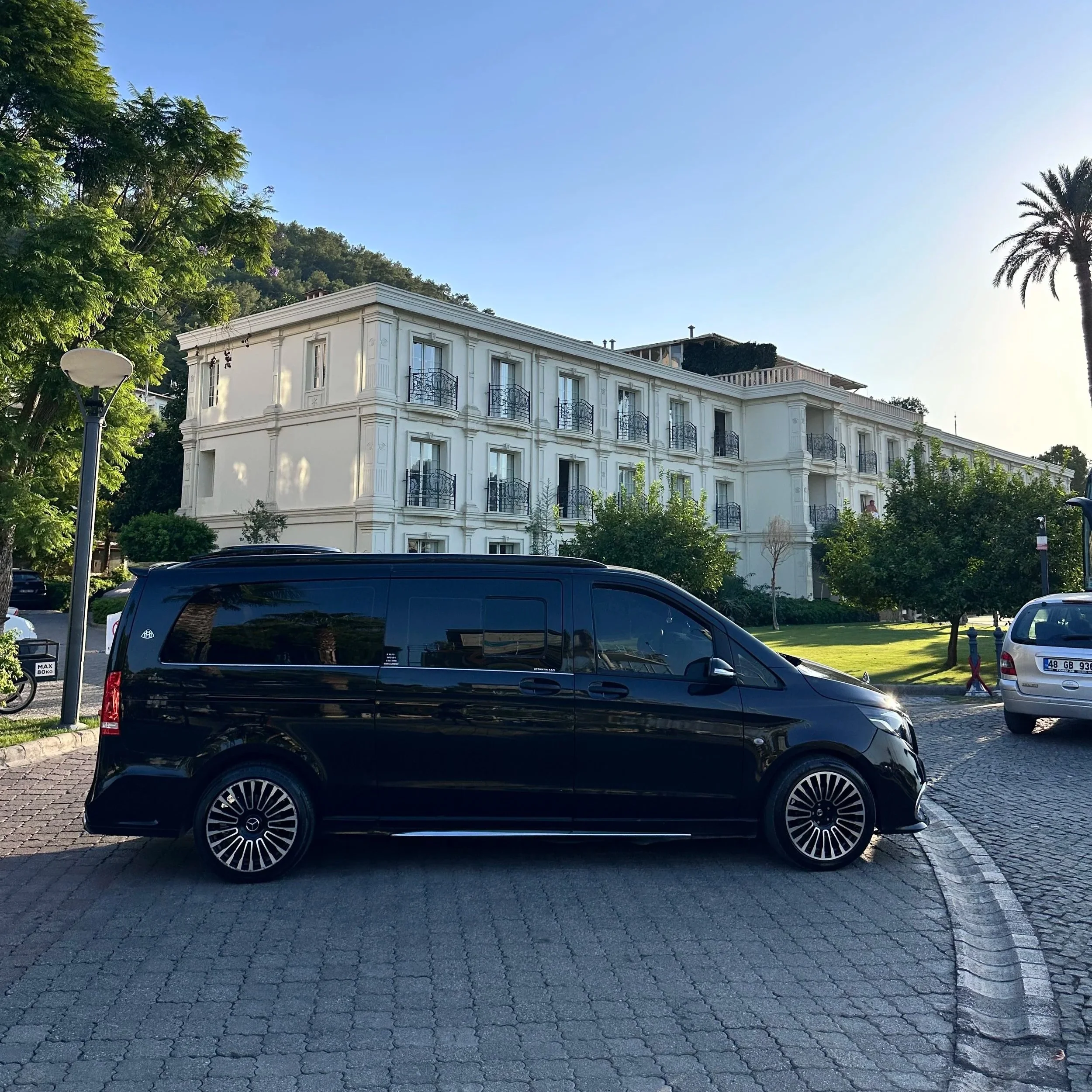 Black van parked on a paved street in front of a white multi-story building with small balconies, surrounded by trees and greenery.