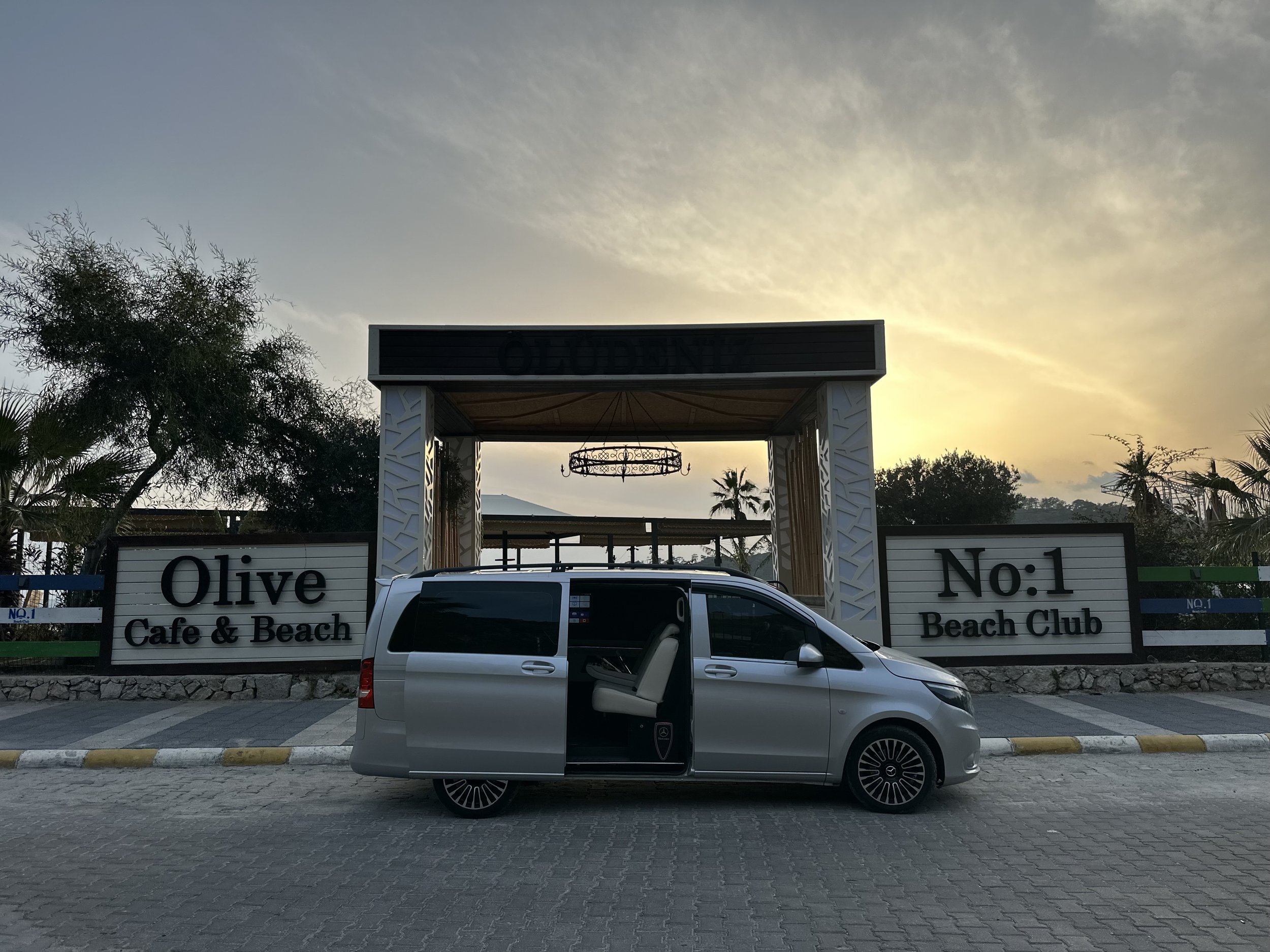 A silver van with an open sliding door parked in front of the entrance to Olive Beach Club at sunset, with trees and a cloudy sky in the background.