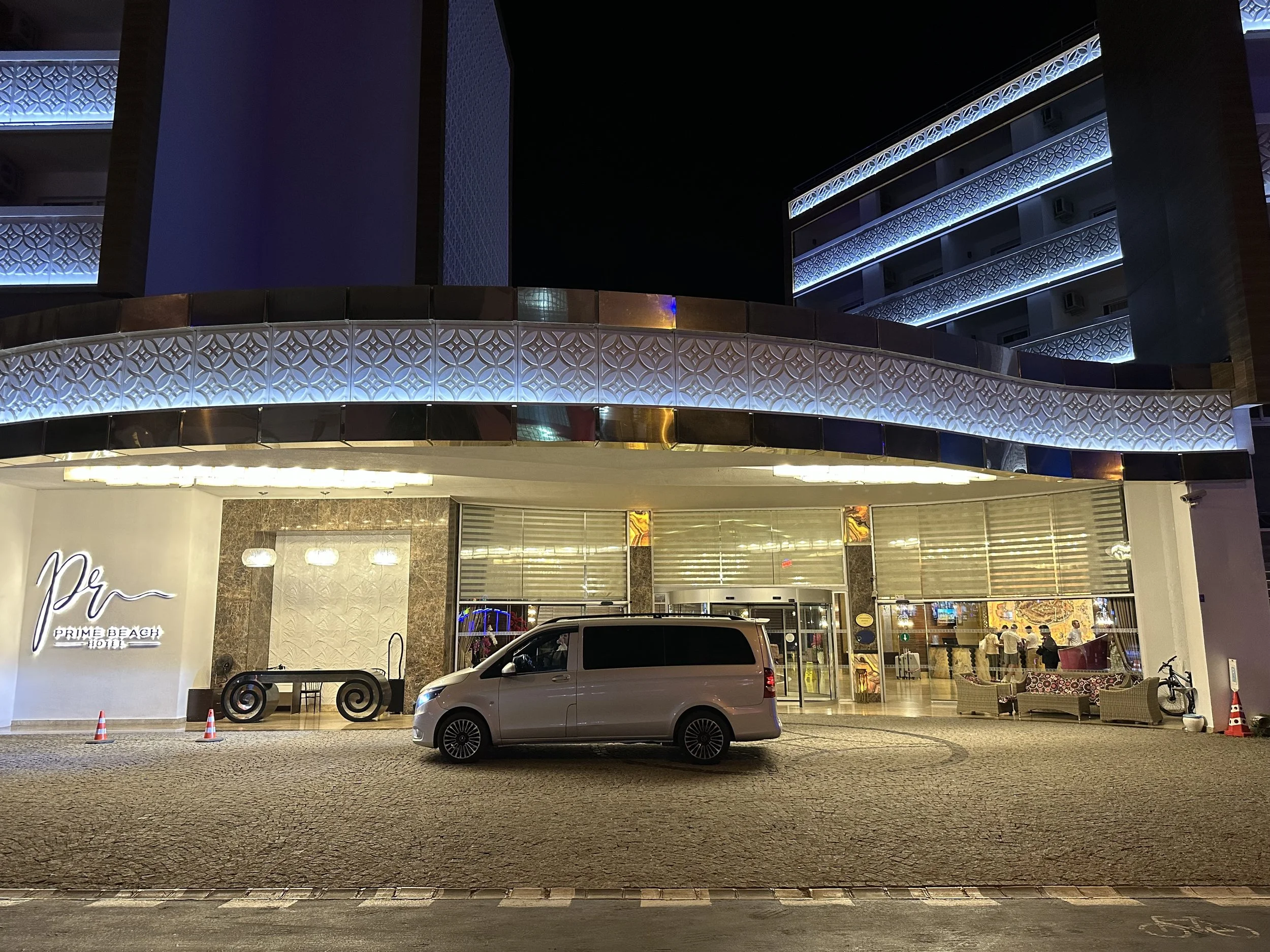 Night view of the entrance to PRIME BEACH HOTEL with a white van parked in front. The hotel has modern architecture with decorative lighting, outdoor furniture including a bench with swirl details, and umbrellas. Glass doors reveal a glimpse of the h