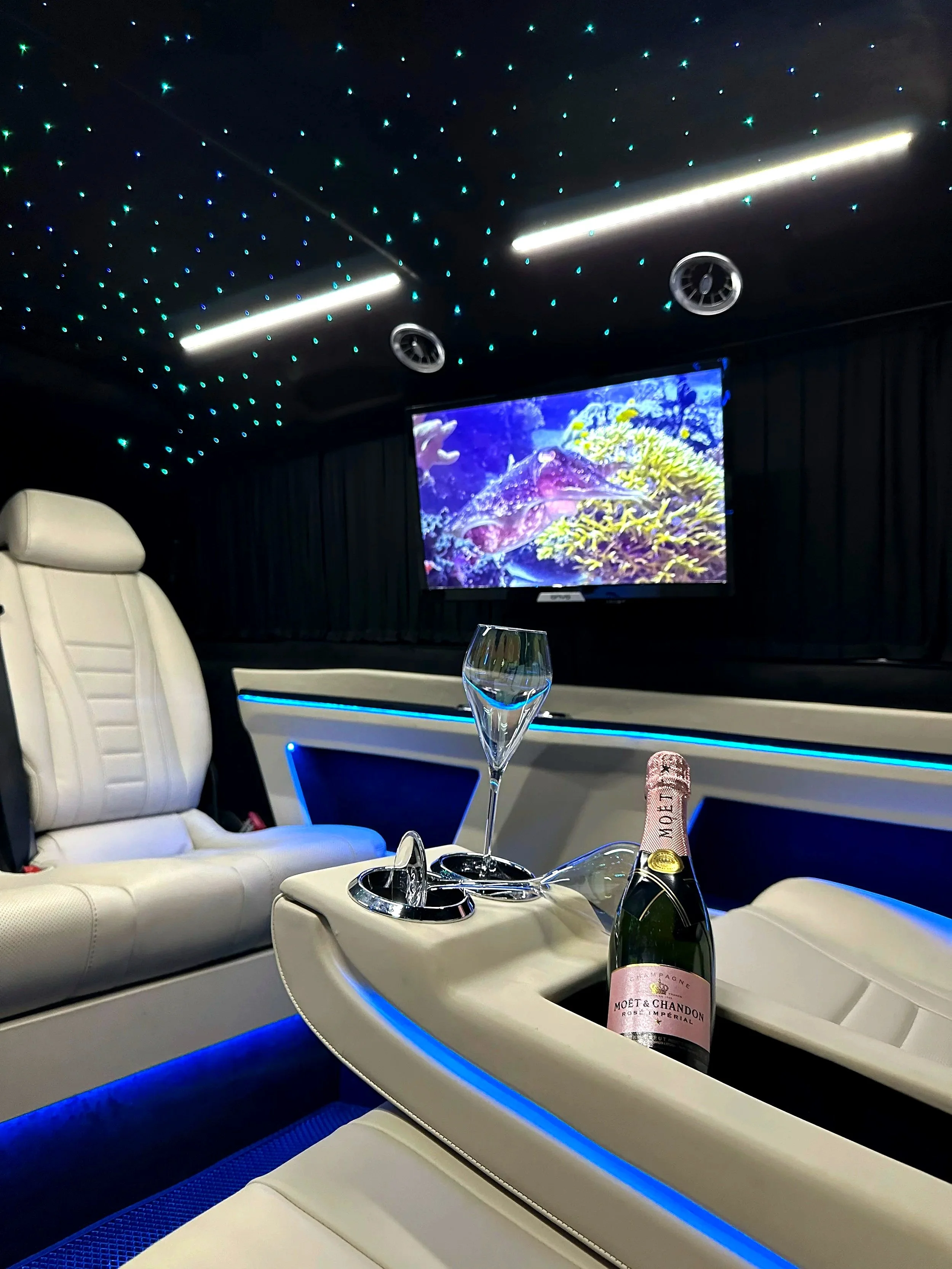 Luxury private entertainment room with a TV showing underwater scene, white leather seats, a bottle of Moët & Chandon champagne, and empty champagne flutes on a beige center console.