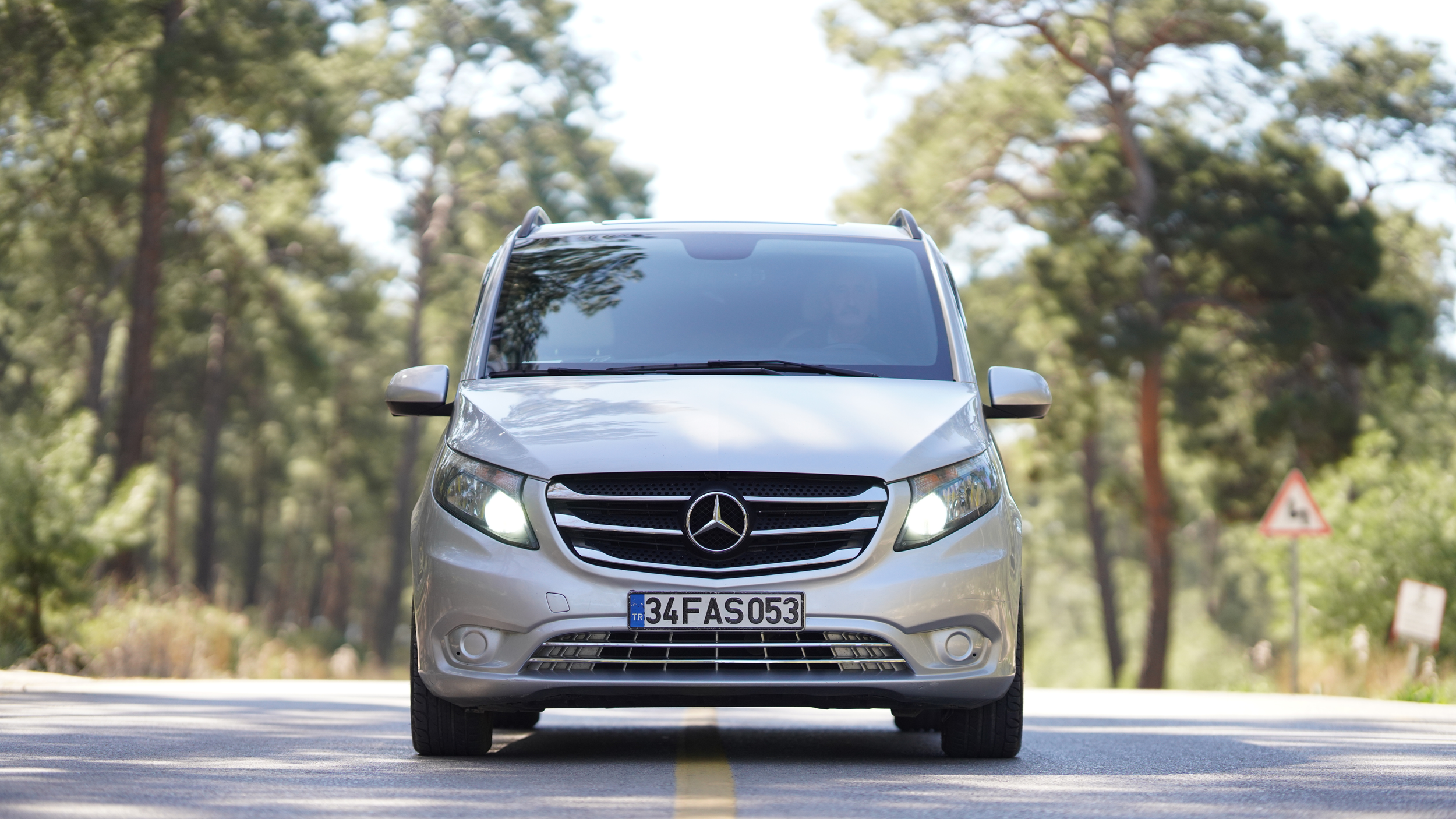 Silver Mercedes-Benz van driving on a forested road, with trees and road signs in the background.