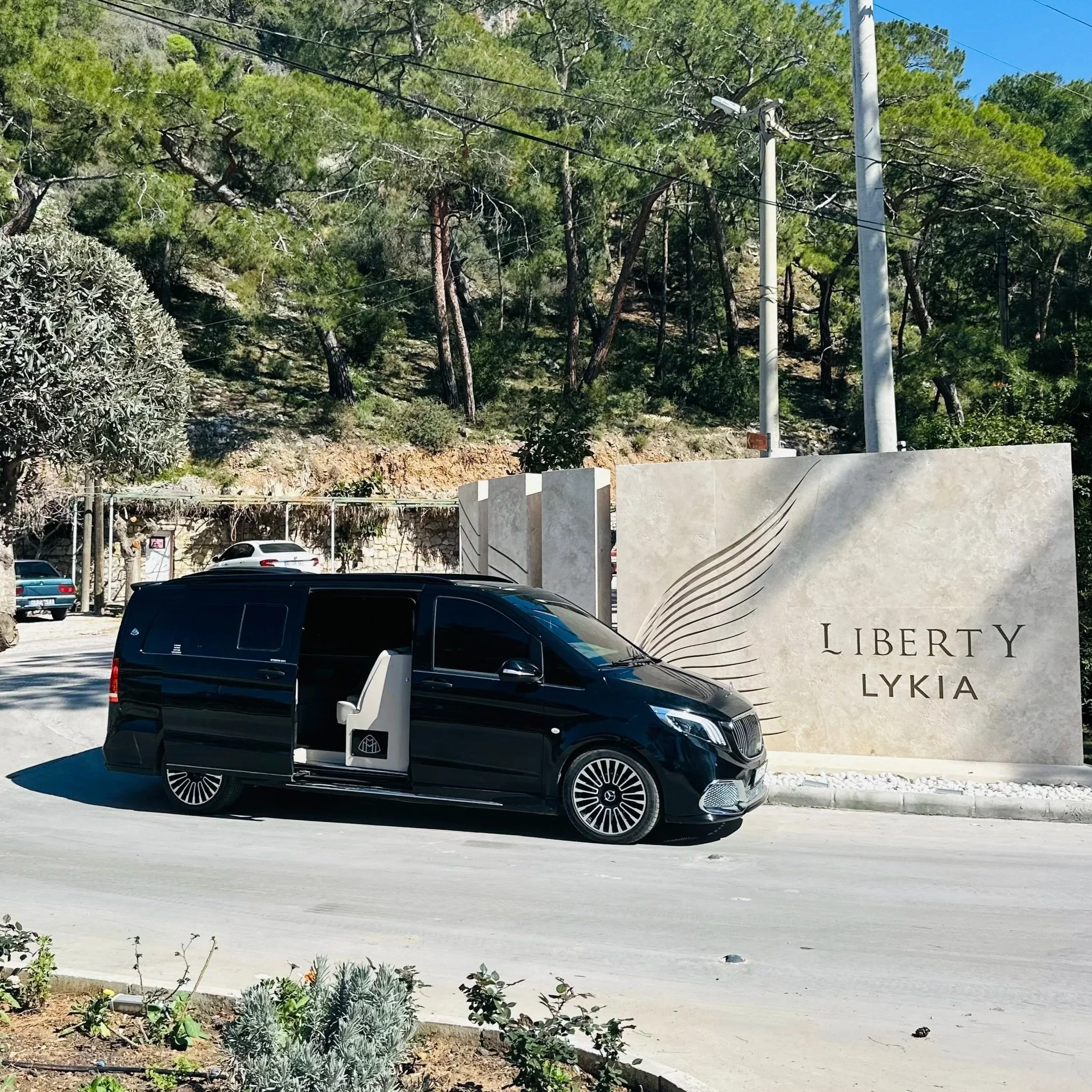 A black van with an open sliding door and a white chair inside, parked in front of a large stone sign for Liberty Lykia with a stylized leaf design, surrounded by a natural landscape of trees on a sunny day.