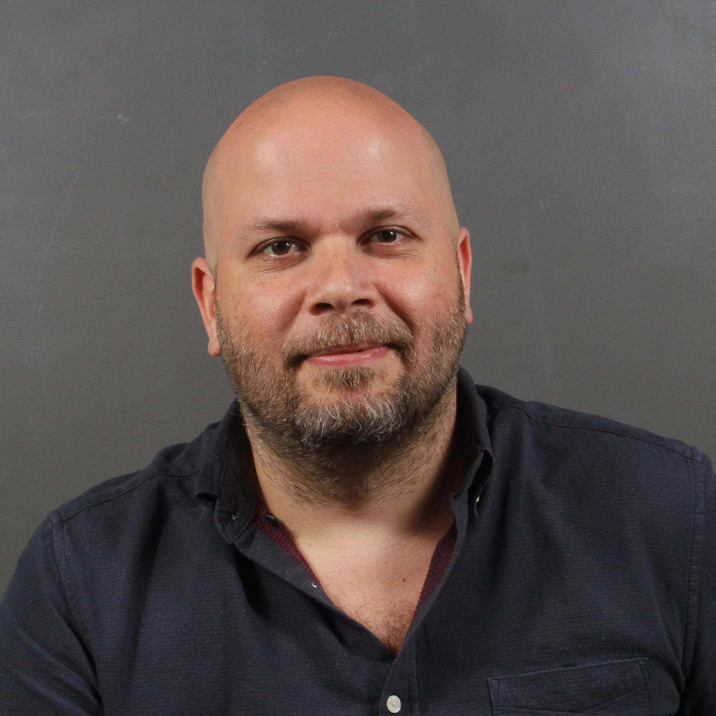 A man with a shaved head and beard in a dark button-up shirt against a gray background.