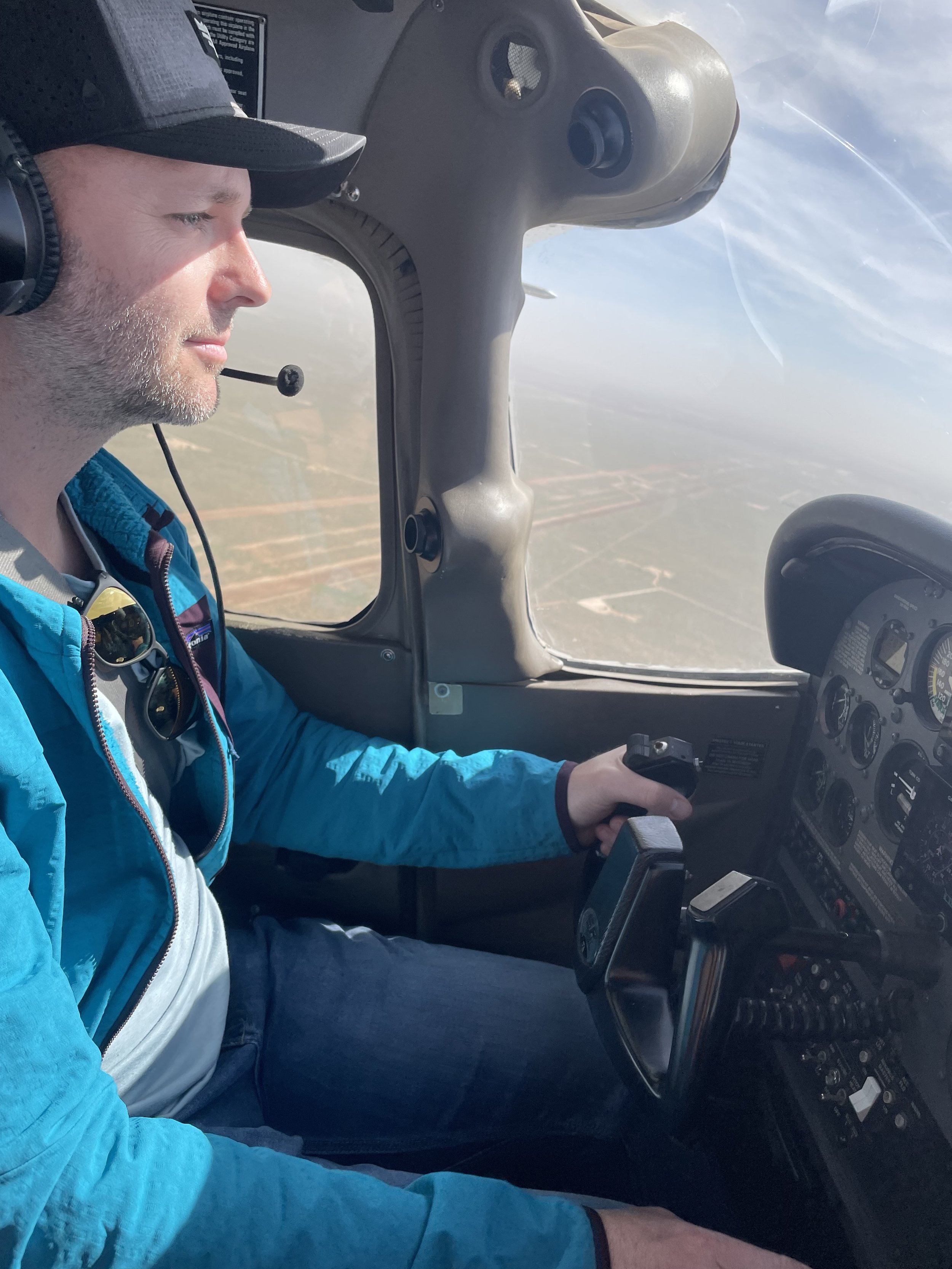 A man wearing a black cap, blue jacket, and headphones flying a small aircraft, looking focused. The cockpit instruments are visible, and a cloudy sky can be seen through the window.