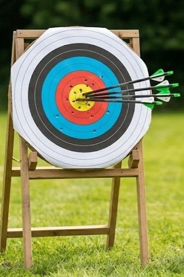 Archery target with multiple arrows shot into the bullseye, set outdoors on a wooden stand.