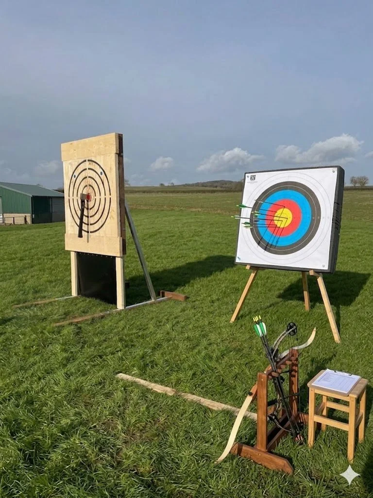 An outdoor archery range with a wooden target on the left and a bullseye target on a stand on the right. Arrows are shot into the bullseye target. A bow and arrow rest with a few arrows are set up on a small stool in the foreground, with a clipboard on top.