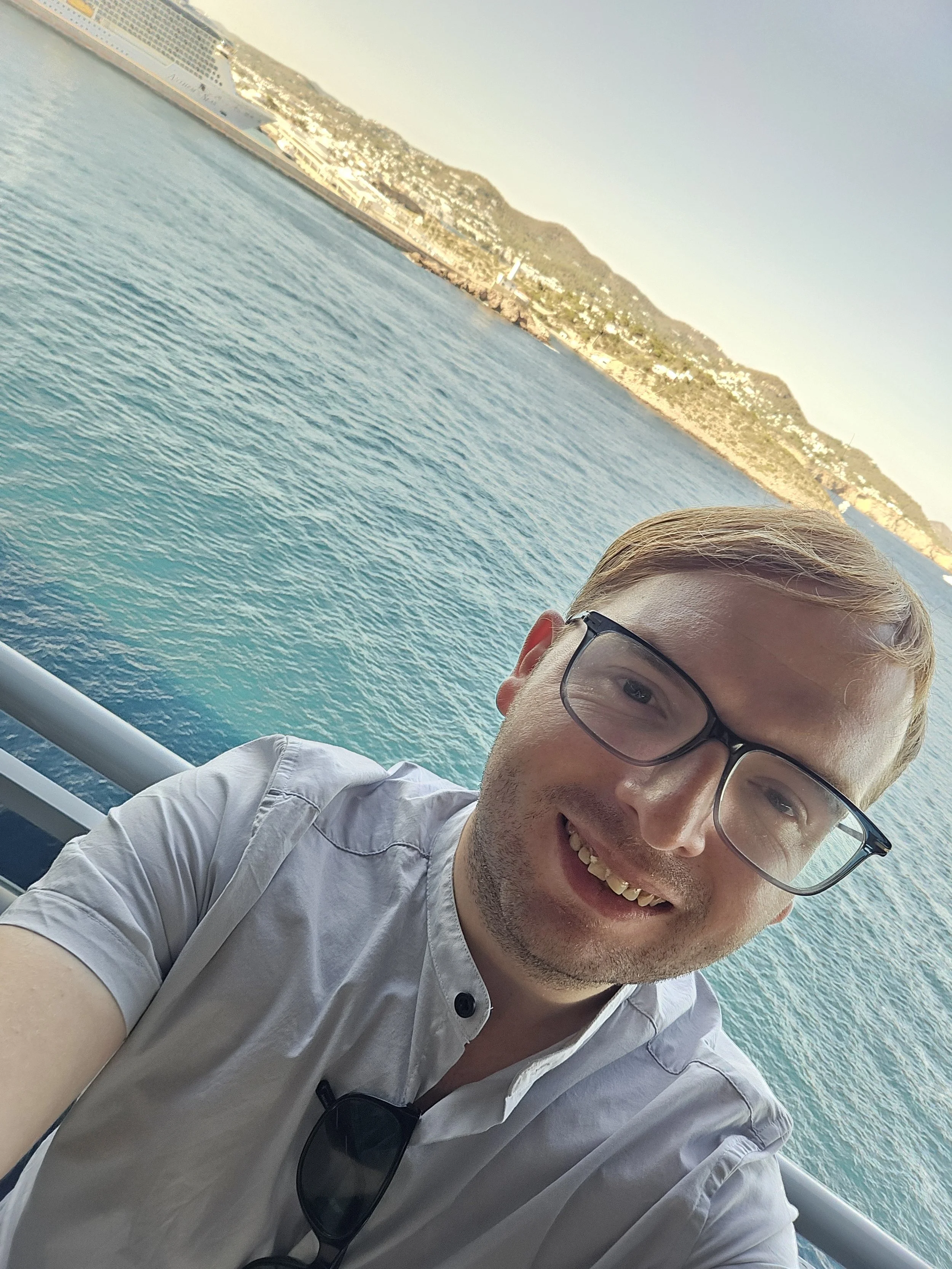 A smiling man with glasses taking a selfie on a boat with a large body of water and a distant shoreline with mountains in the background.