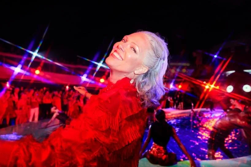 A smiling woman with gray hair, wearing a red outfit, at a vibrant party with colorful lights and people in the background.