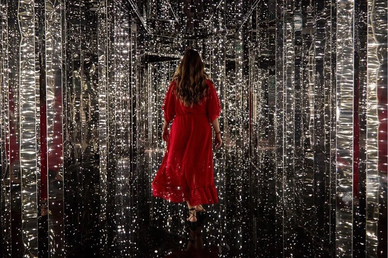 A woman in a red dress walking through a mirrored tunnel decorated with numerous small, bright lights.