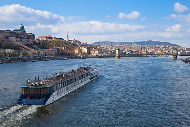 A large river with a cruise boat sailing, city buildings on the hills, and a bridge in the background under a partly cloudy sky.