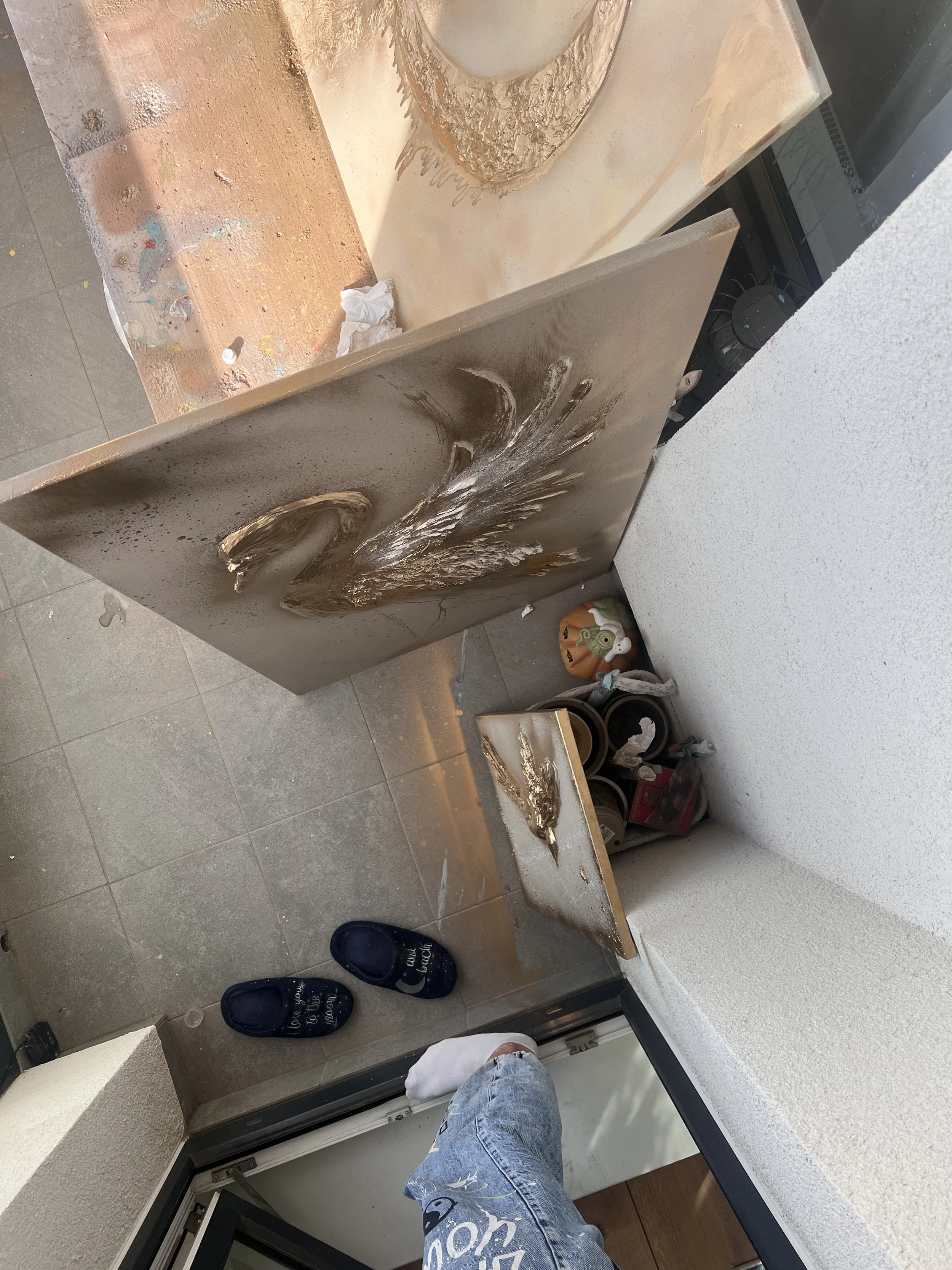 Nedokončené umělecké dílo se zlatou barvou na plátně a socha v pozadí, umístěné na podlaze poblíž polic s výtvarnými potřebami v ateliéru.
Jak žijí umělci