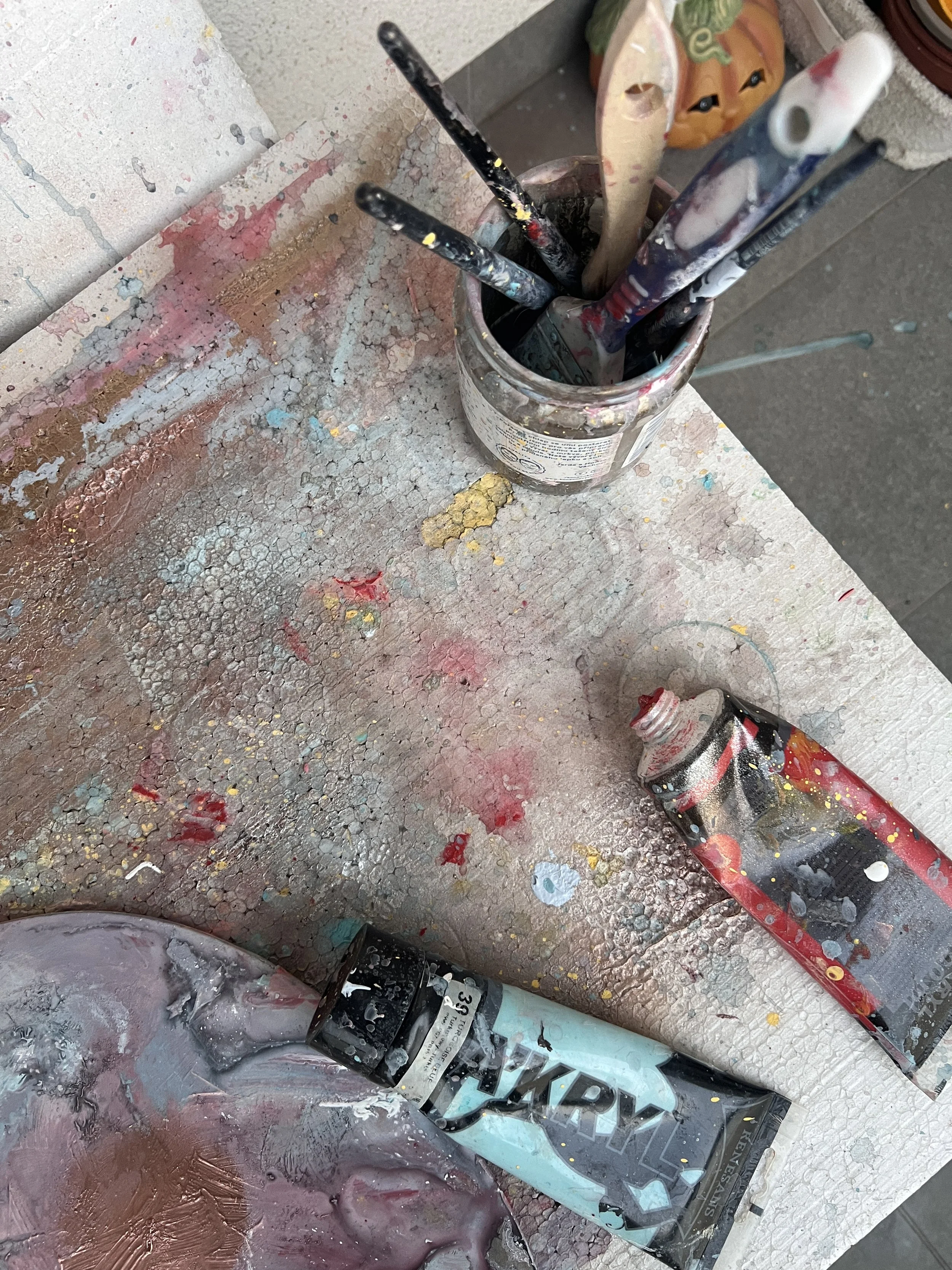 An art workspace with paint splatters and tools, including brushes in a jar, and tubes of paint on a textured, paint-covered surface.