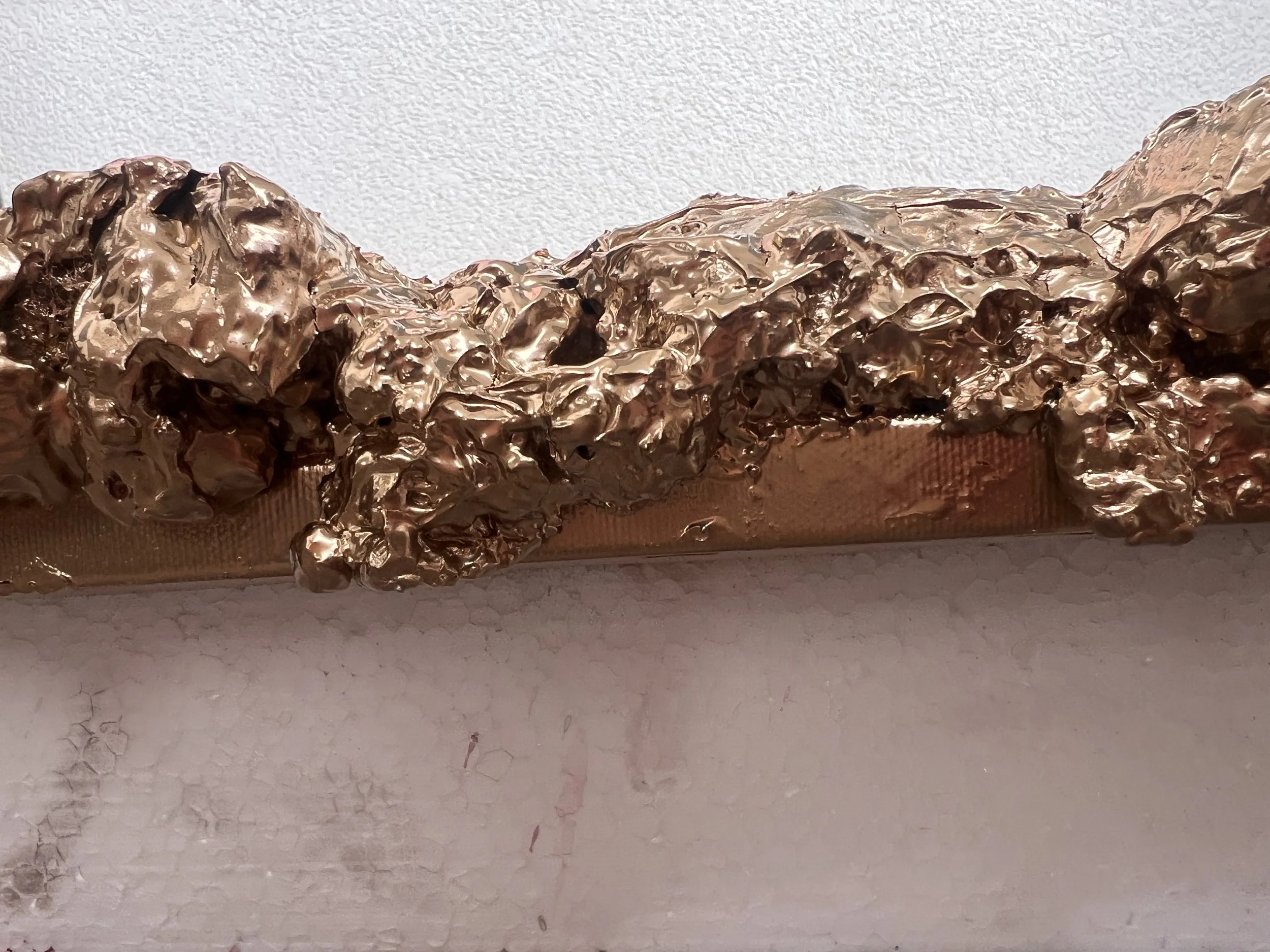 Velká, nepravidelně tvarovaná socha vytvořená z pomačkaného a vrstveného kovového materiálu v zlaté nebo bronzové barvě, umístěná na jednoduchém bílém pozadí, ve stylu fine art. Ideální pro milovníky moderního umění, abstraktních a textur