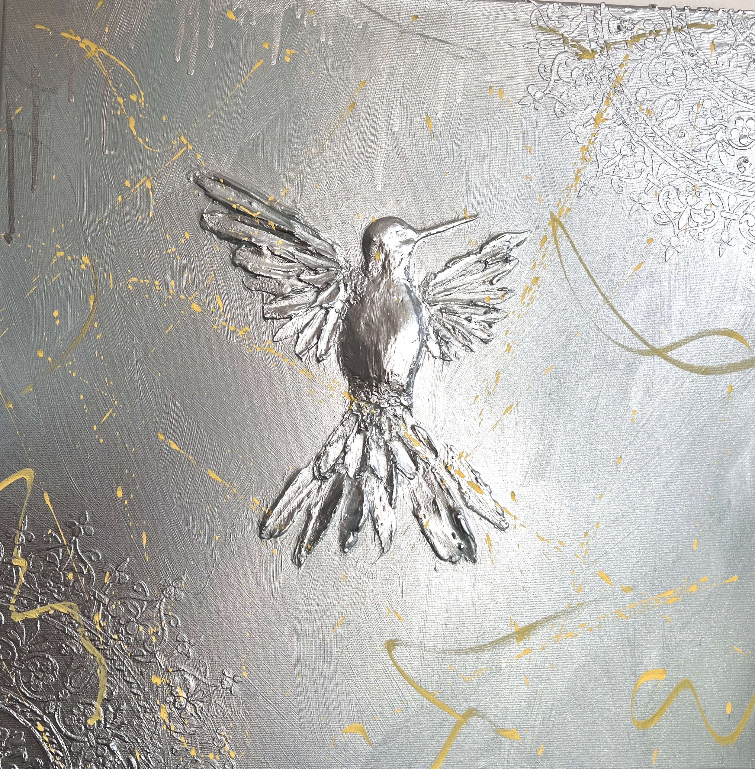 A metallic wall art featuring a raised silver hummingbird with outstretched wings, surrounded by gold decorative splatters and intricate ornate patterns in the background.