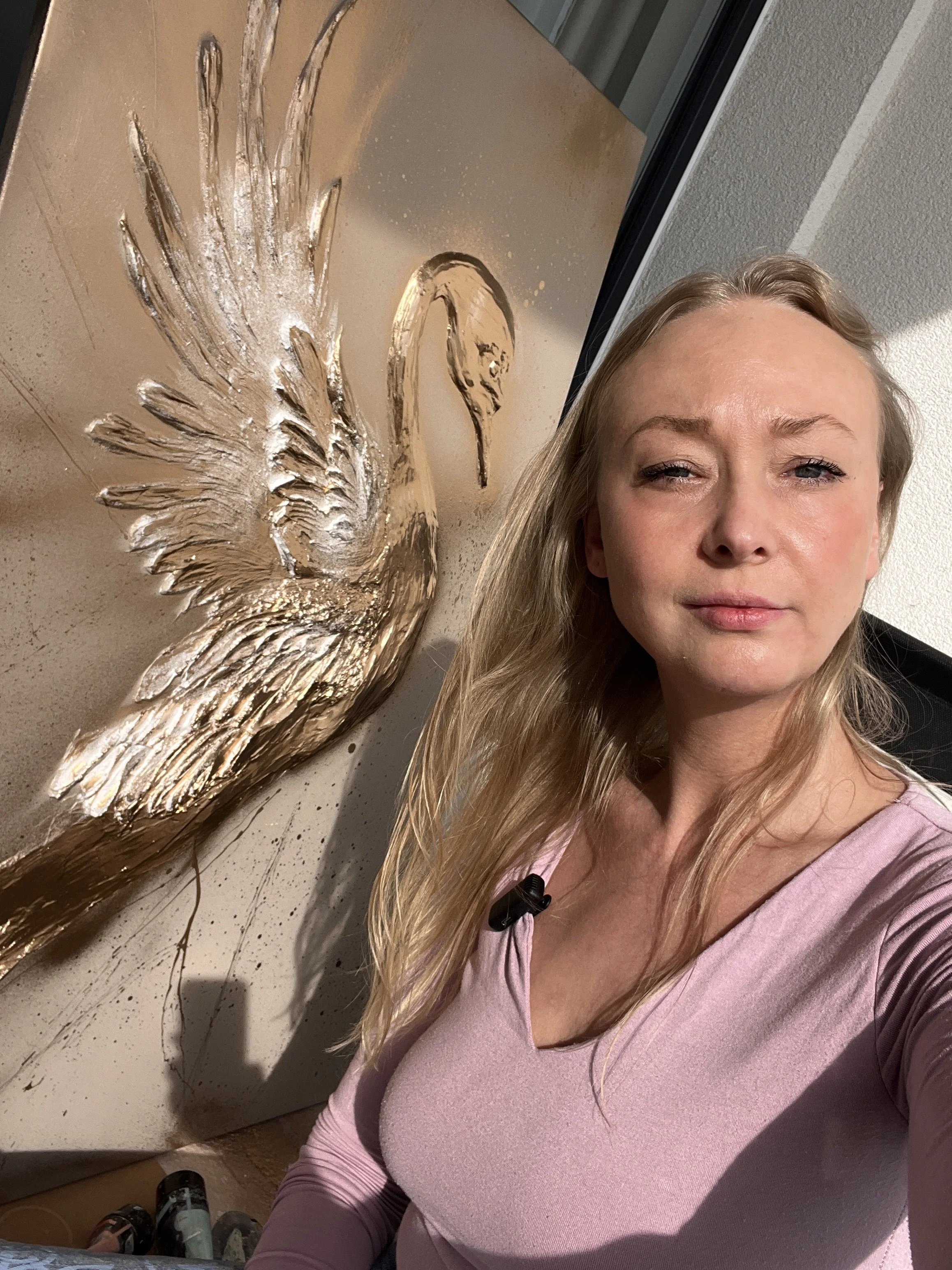 Žena s dlouhými blond vlasy si pořizuje selfie vedle dekorativní zlaté kovové sochy labutě na stěně. Ideální pro milovníky umění, interiérových dekorací a moderních wall art designů.