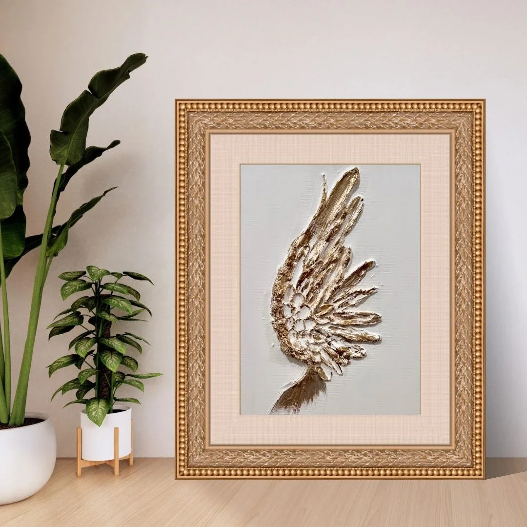 Decorative framed artwork of a metallic winged figure on a white background, placed on a wooden surface alongside potted plants.