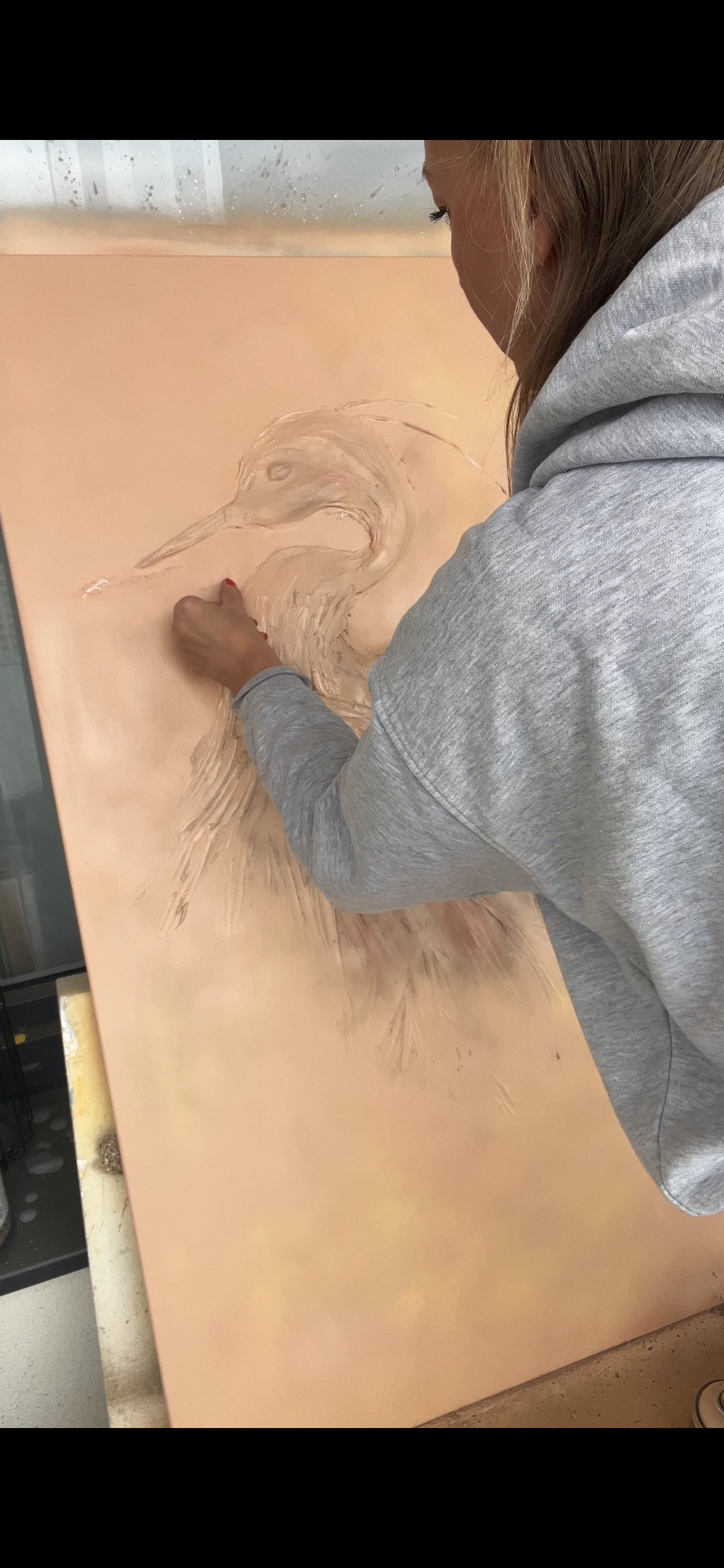 Umělec v šedé mikině kreslí realistický portrét ptáka na velké béžové plátno pomocí křídy nebo pastelu.