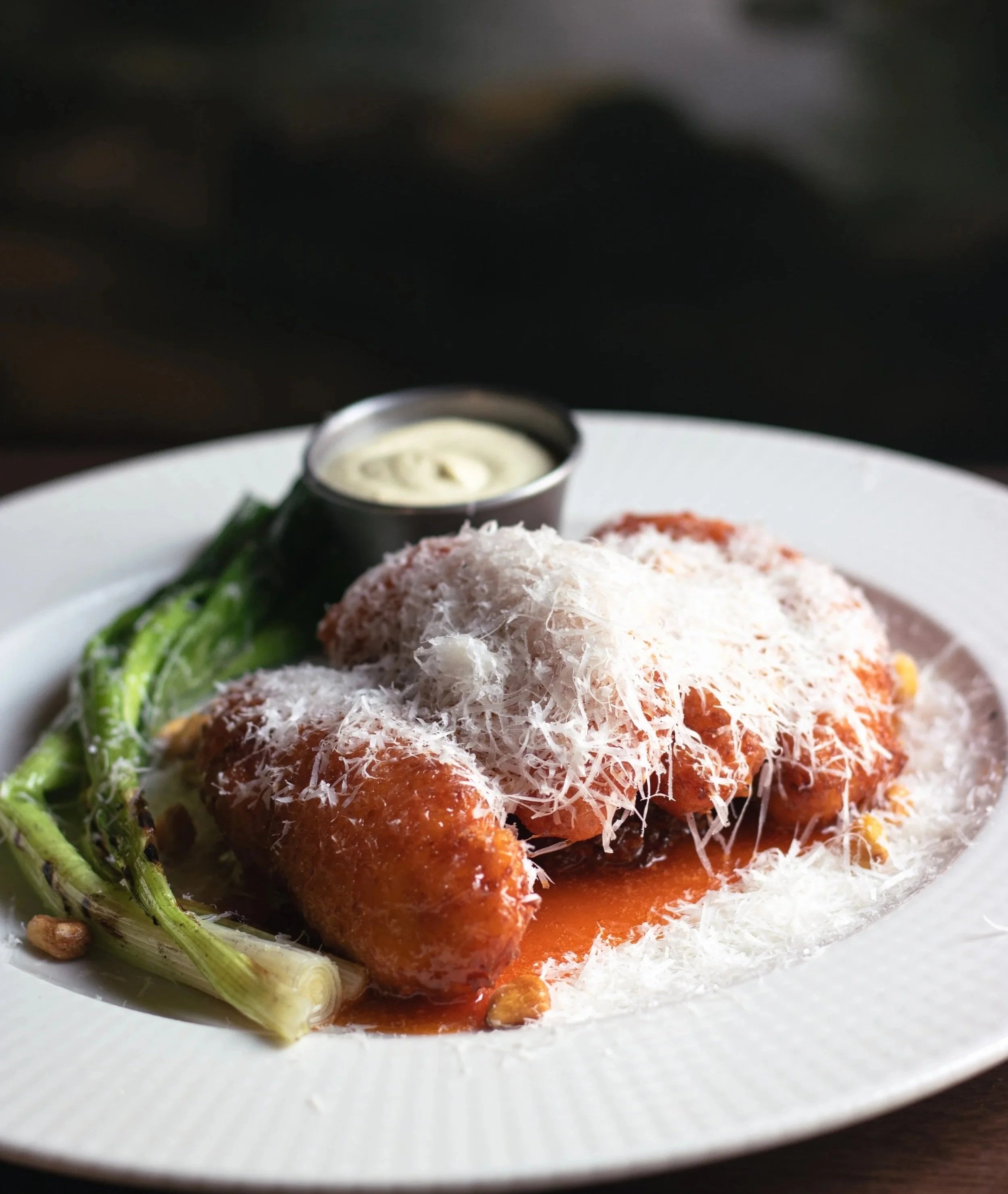 Close-up of a plate with breaded fried food topped with shredded cheese, served with green vegetables, red sauce, and a small container of creamy sauce, on a white plate.