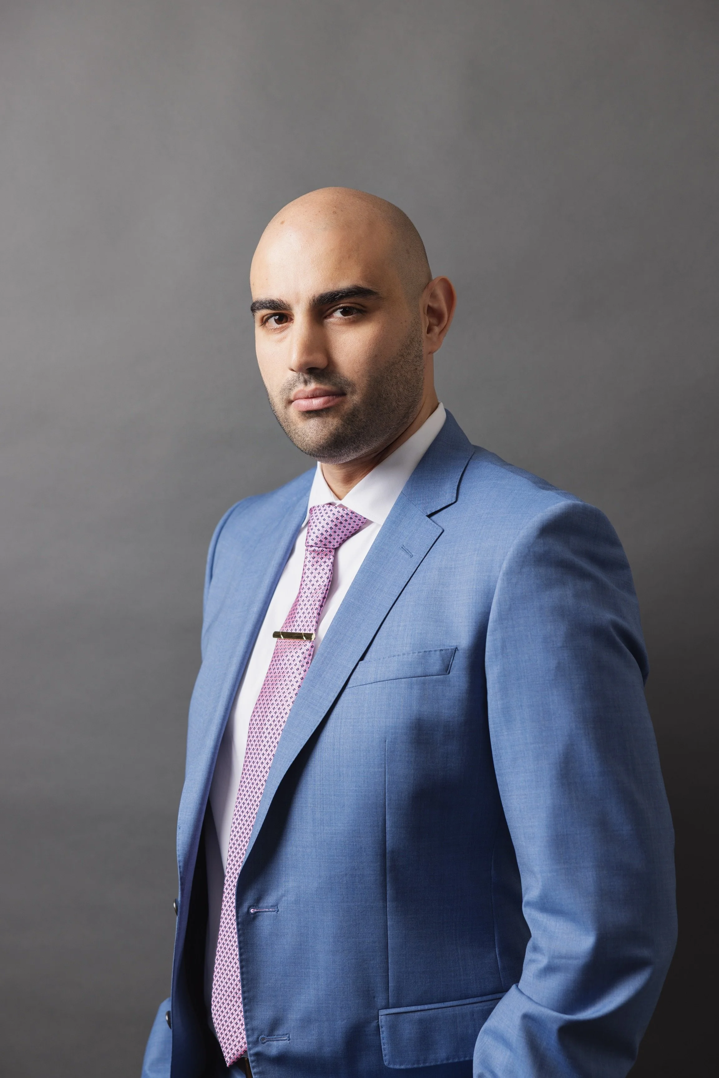 Portrait of a bald man wearing a blue suit, light pink tie, and white shirt against a gray background.