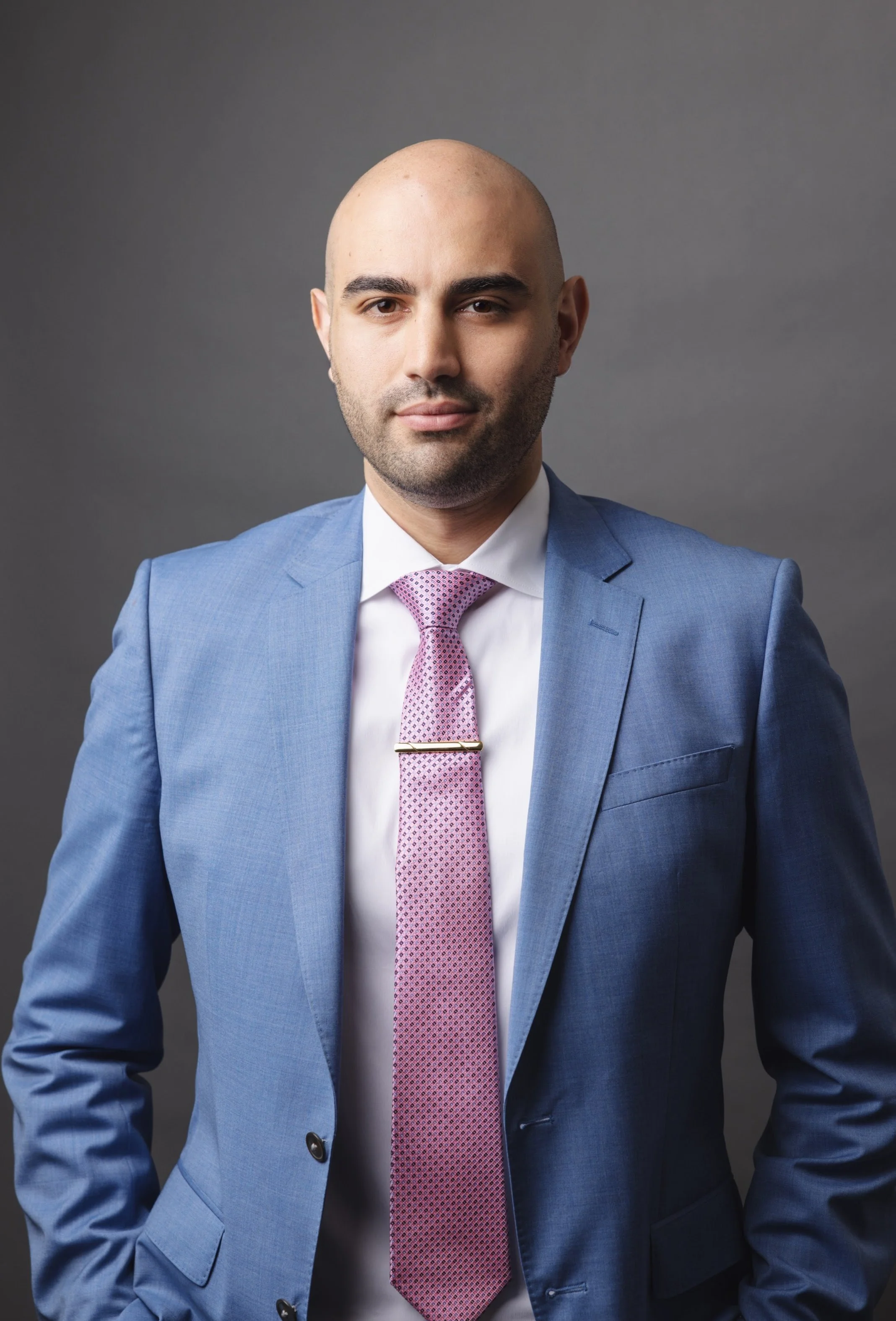 Professional man wearing a blue suit, pink tie, and white shirt against a gray background.