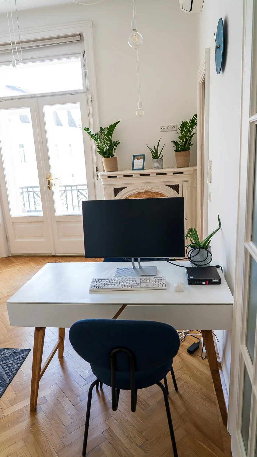 Ein Home-Office mit weißem Schreibtisch, einem schwarzen Monitor, Tastatur, Maus, einem kleinen Drucker, einer Zimmerpflanze, einem blauen Stuhl, weißen Wänden, einem Kamin mit Pflanzen, einer Uhr an der Wand und einem Balkon im Hintergrund.