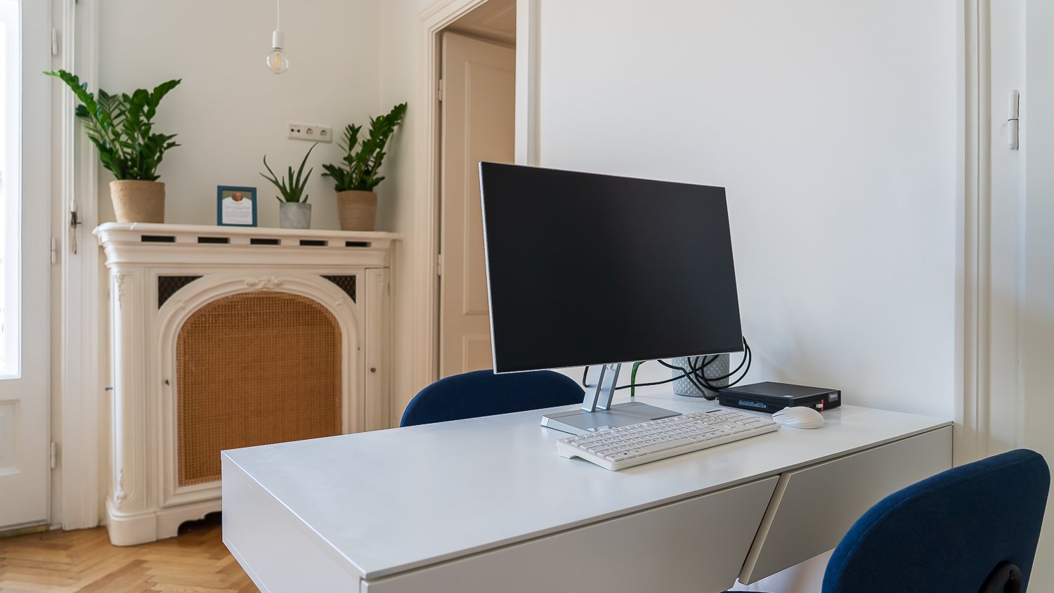 Ein moderner Arbeitsbereich mit einem weißen Schreibtisch, einem schwarzen Computermonitor, einer Tastatur und einer Maus. Im Hintergrund eine weiße Wand mit einer Kaminatrappe, darauf drei Pflanzen in Töpfen, und ein offener Türrahmen.
