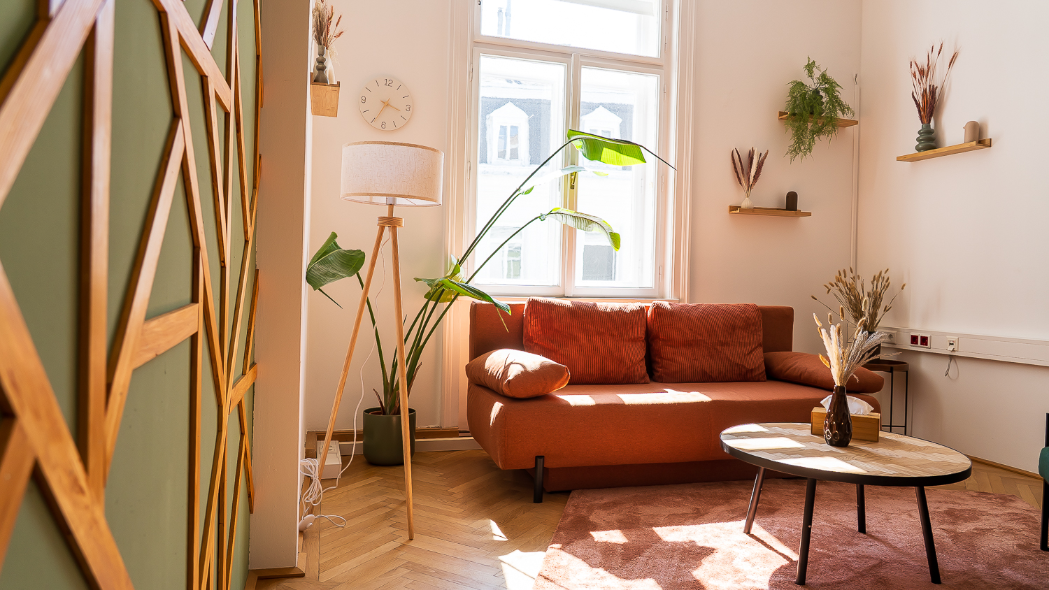 Helles Wohnzimmer mit Sofa, Couchtisch, Topfpflanzen, Wandregalen und Sonnenlicht.