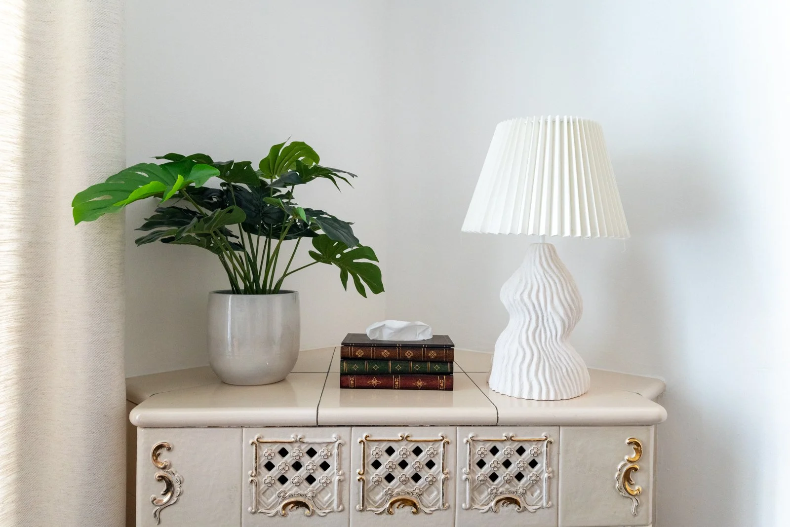 Ein weißer Sideboard mit dekorativen Schnitzereien, auf dem sich ein großer grüner Pflanzentopf mit Monstera-Pflanze, eine weiße Tischlampe mit gewelltem Design und ein Stapel alter Bücher mit einem Papiertaschentuch befinden.