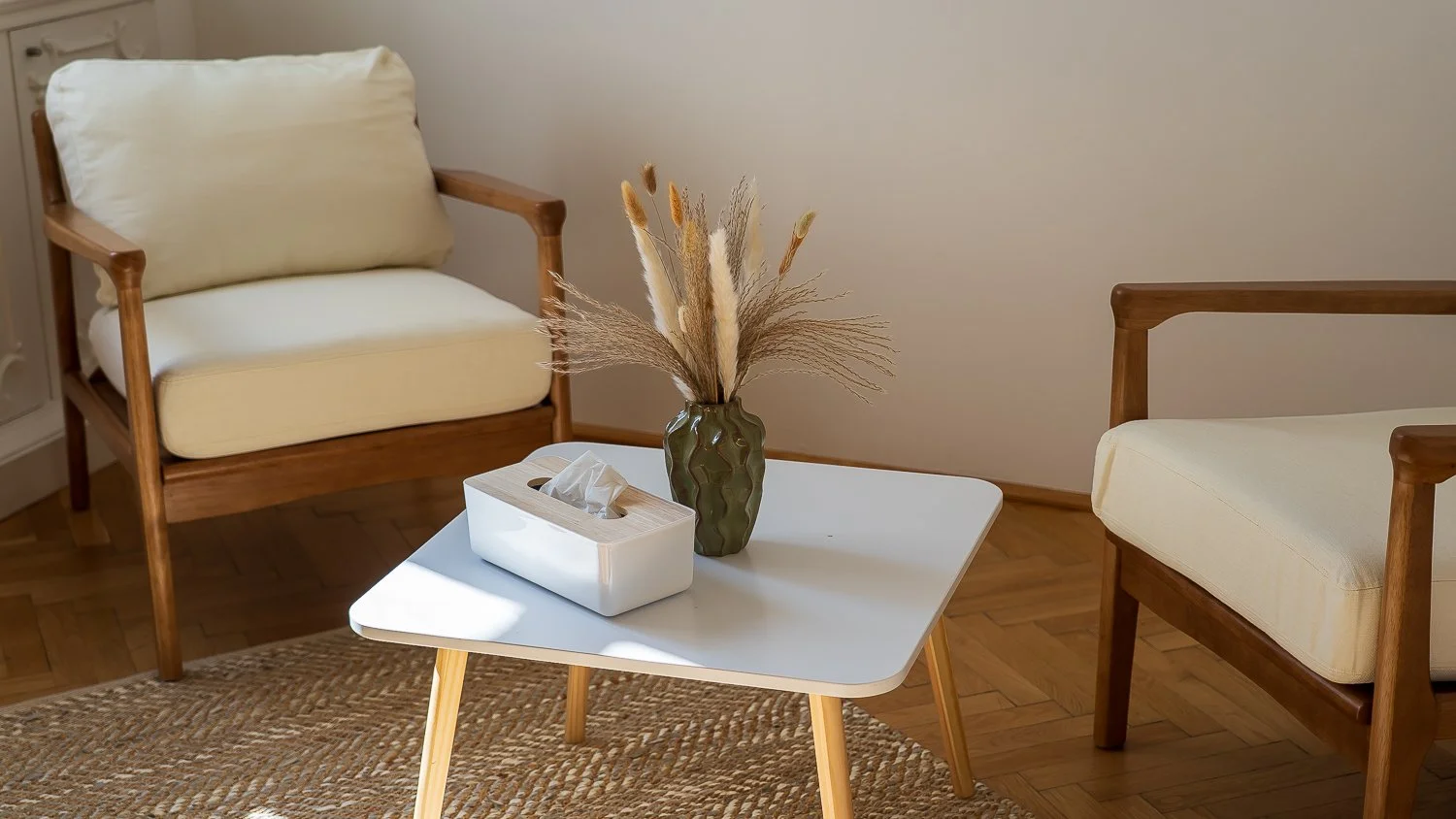 Ein Wohnzimmer mit zwei Holzsesseln mit cremefarbenen Kissen, einem weißen Couchtisch mit einer Vase mit getrockneten Blumen und einer Schachtel mit Taschentüchern, auf einem geflochtenen Teppich auf Holzboden.