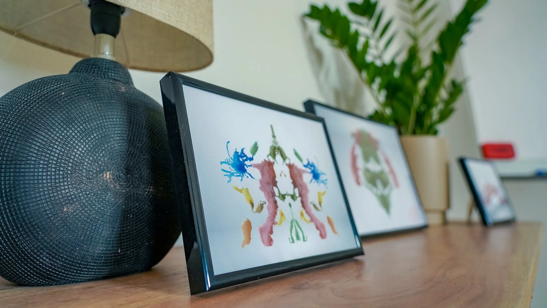 Drei Bilderrahmen mit abstrakten, farbenfrohen Gemälden, eine schwarze dekorative Vase, eine Tischlampe mit einem beigen Lampenschirm, eine grüne Pflanze in einem beige Topf, ein rotes Objekt auf der Wand im Hintergrund auf einer Holzoberfläche