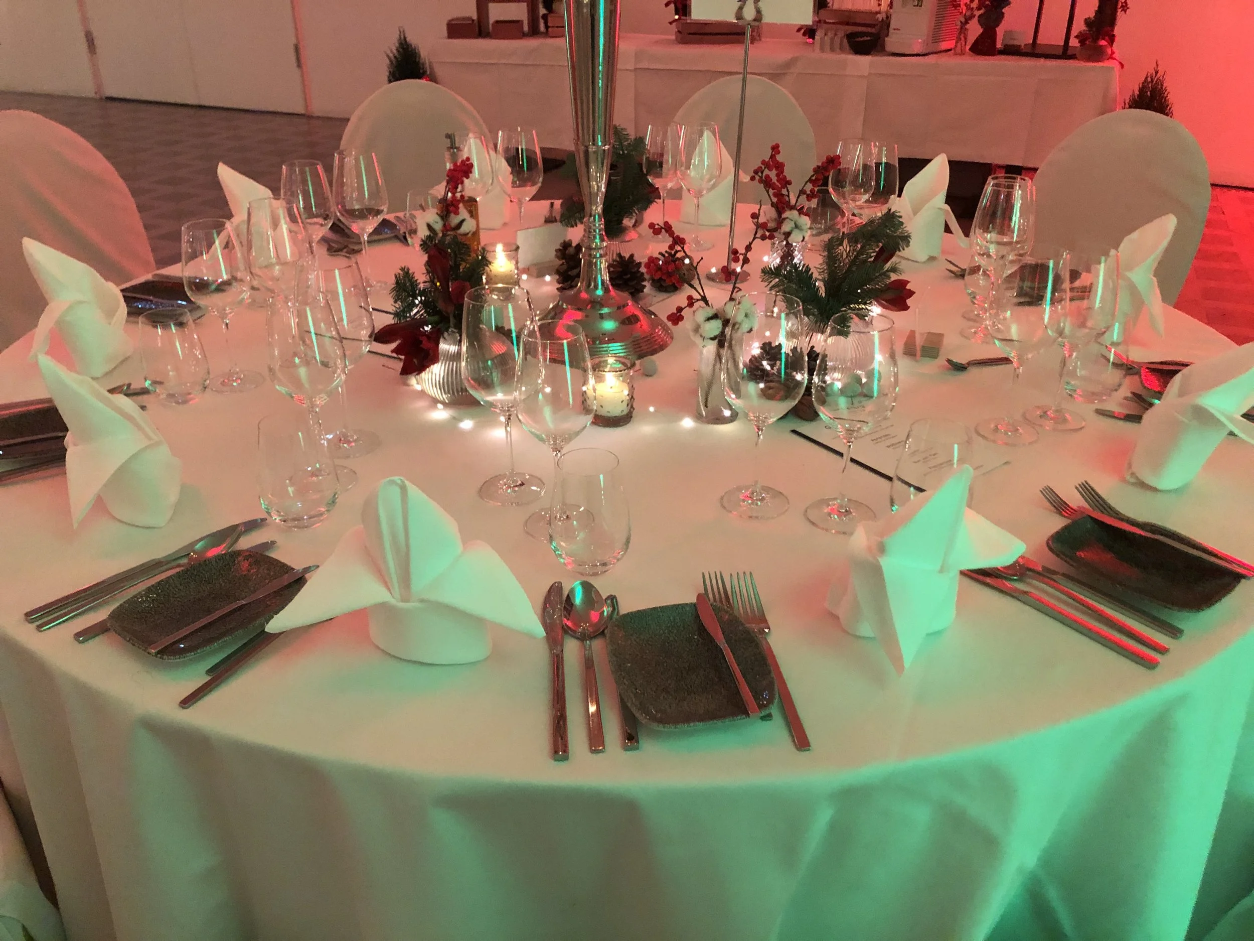 Festlich gedeckter runder Tisch mit weißen Servietten, Gläsern, Kerzen und Blumenschmuck in rotem und grünem Licht.