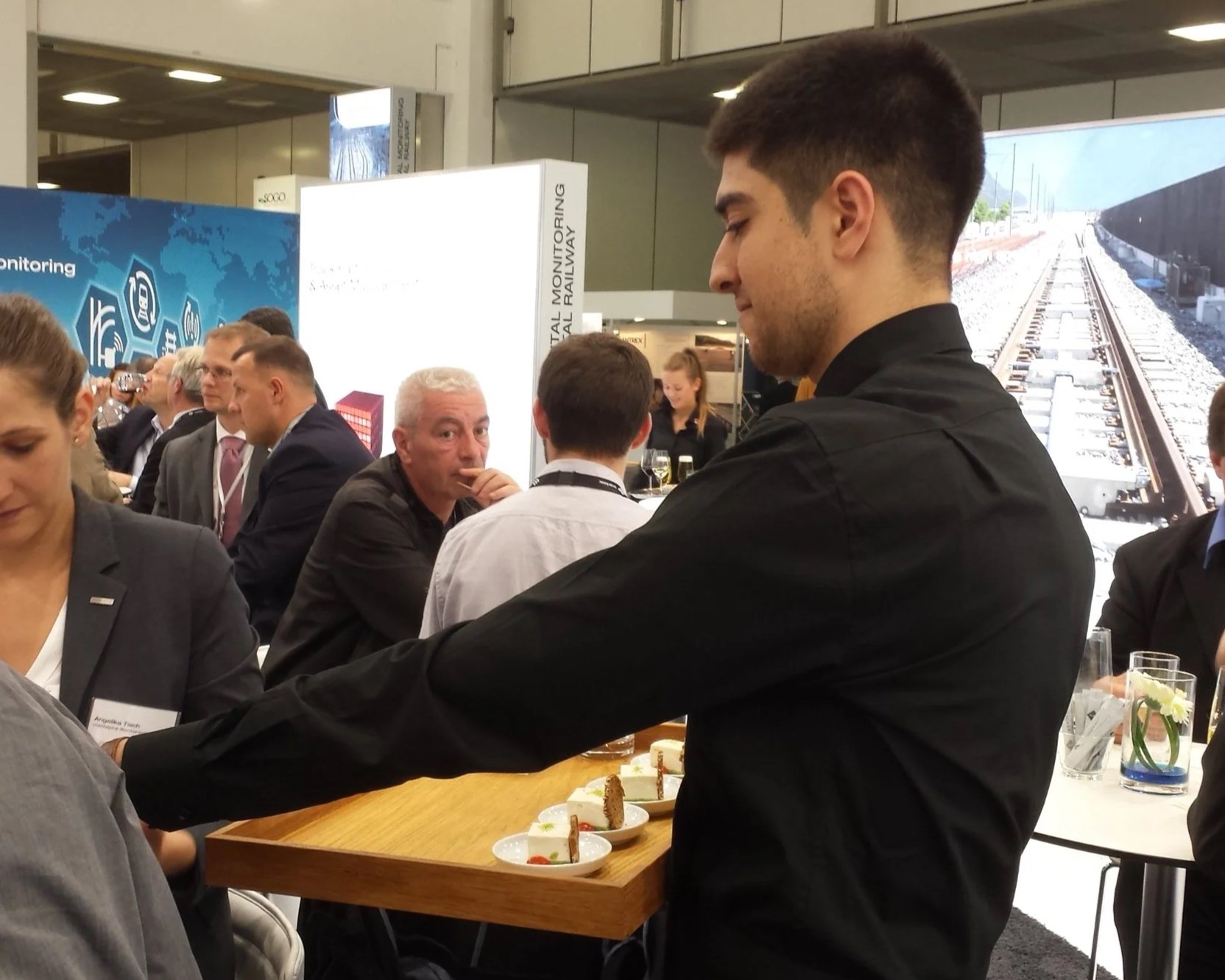 Ein Servicemitarbeiter reicht Fingerfood auf einem Holztablett zwischen Gästen an einem Messestand.