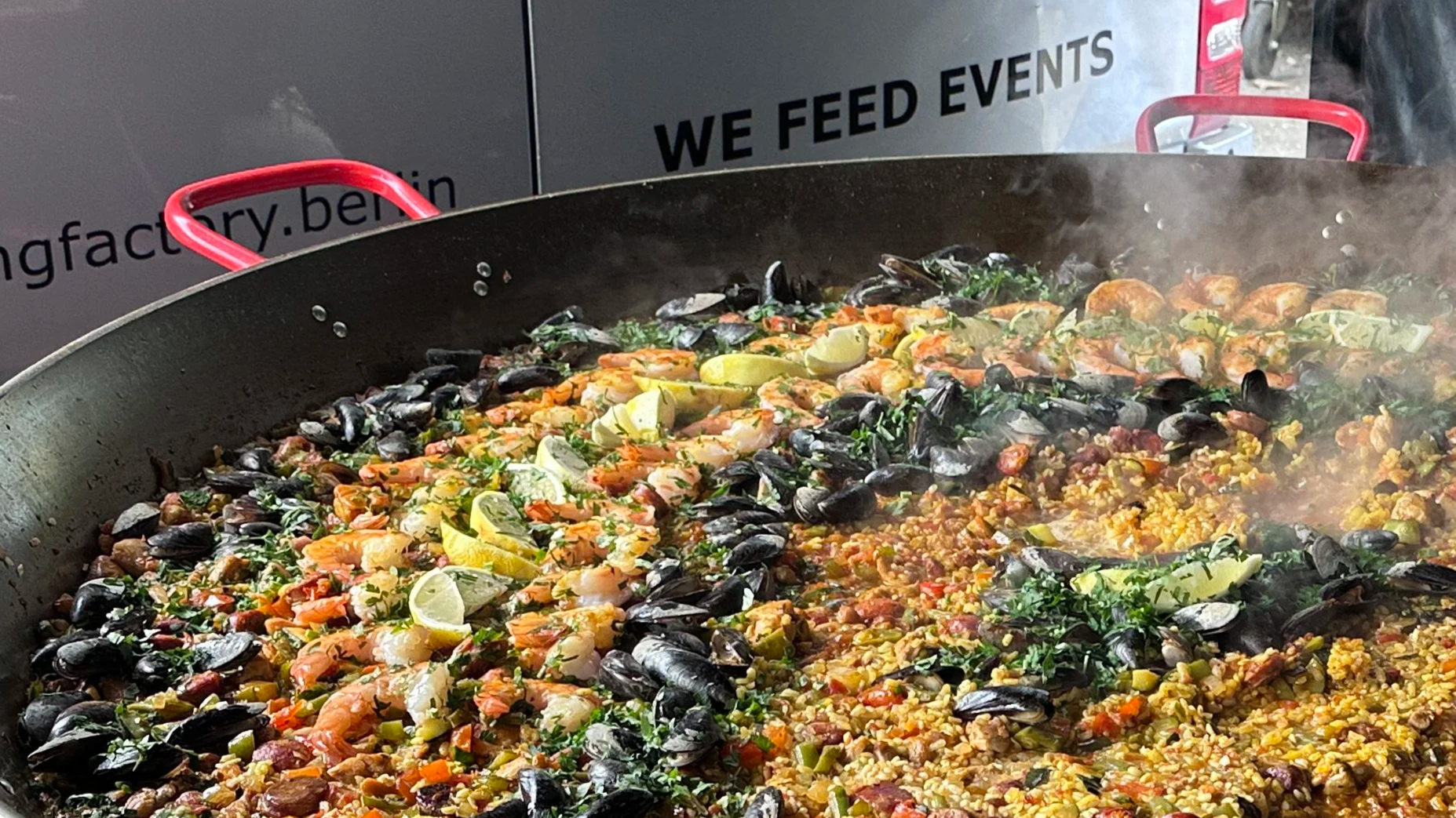 Paella mit Muscheln, Gemüse und Zitronenspalten in einer Riesenpfanne.