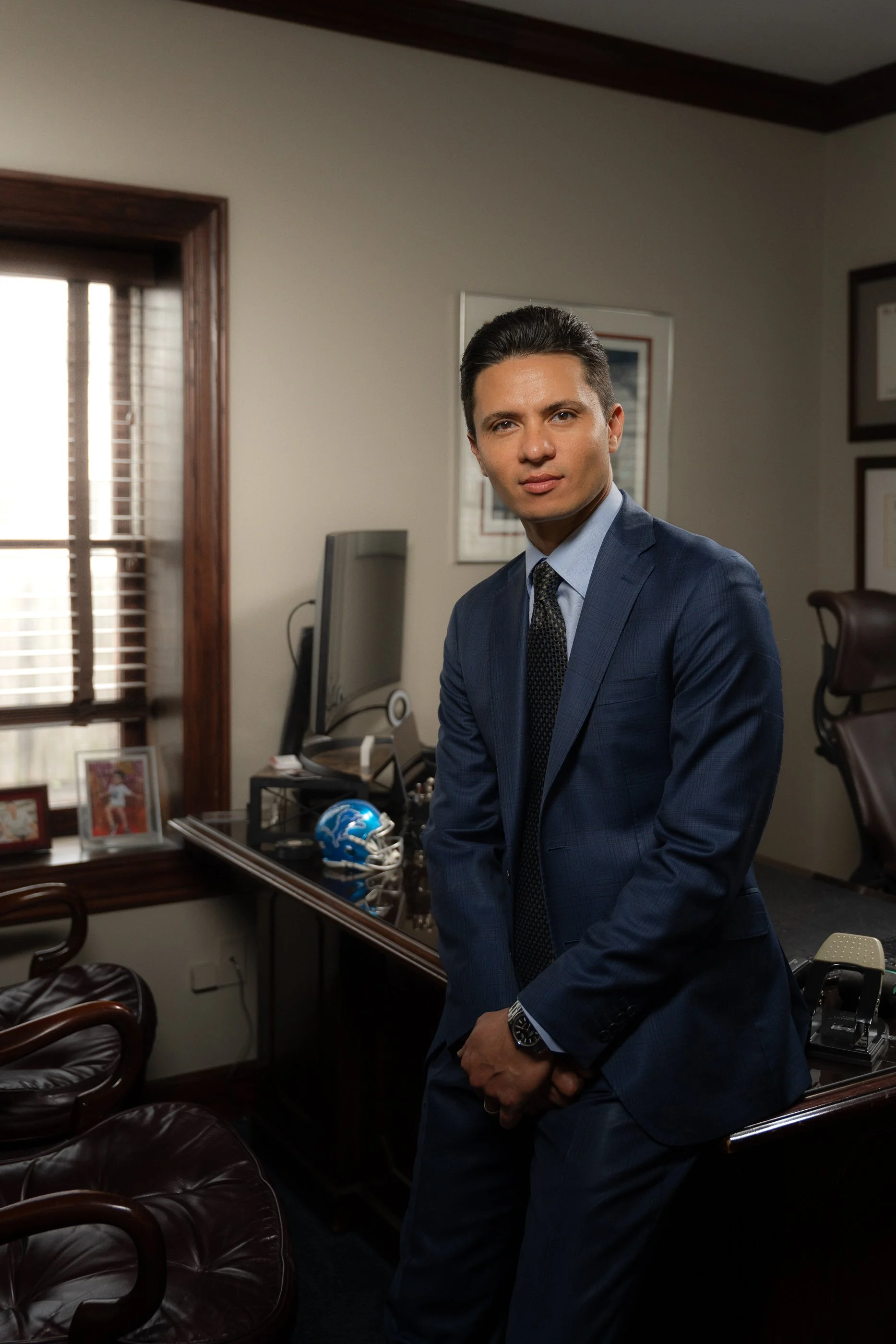 A man in a blue suit with a patterned tie and a watch on his left wrist is standing in an office, looking at the camera. The office has a desk with a blue football helmet, framed photos, and various items. There is a window with wooden trim and blinds behind him, and framed certificates or artwork on the wall.