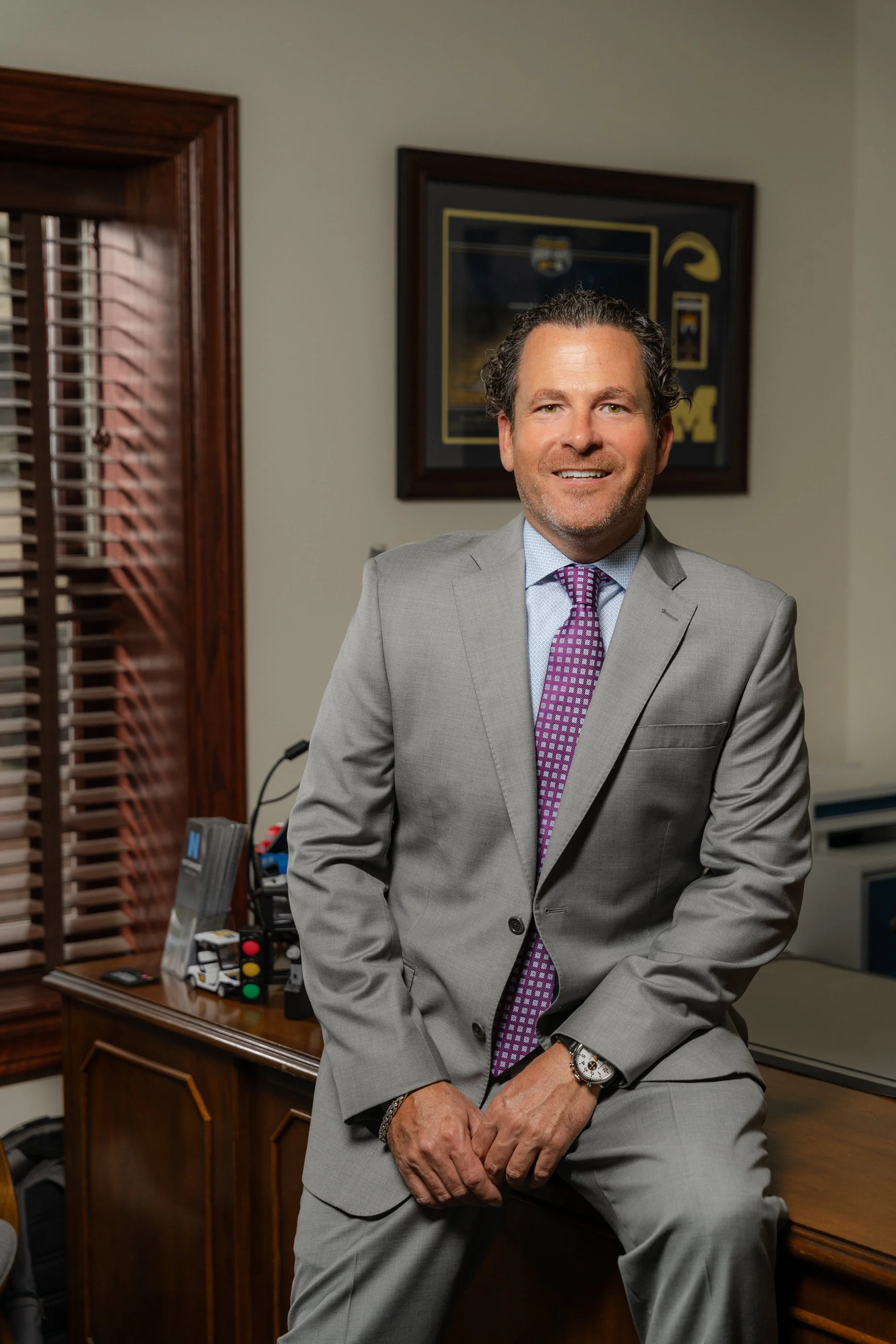 A smiling man in a light gray suit with a purple patterned tie, sitting and leaning against a wooden desk in an office. There is a framed picture on the wall behind him and office supplies on the desk.