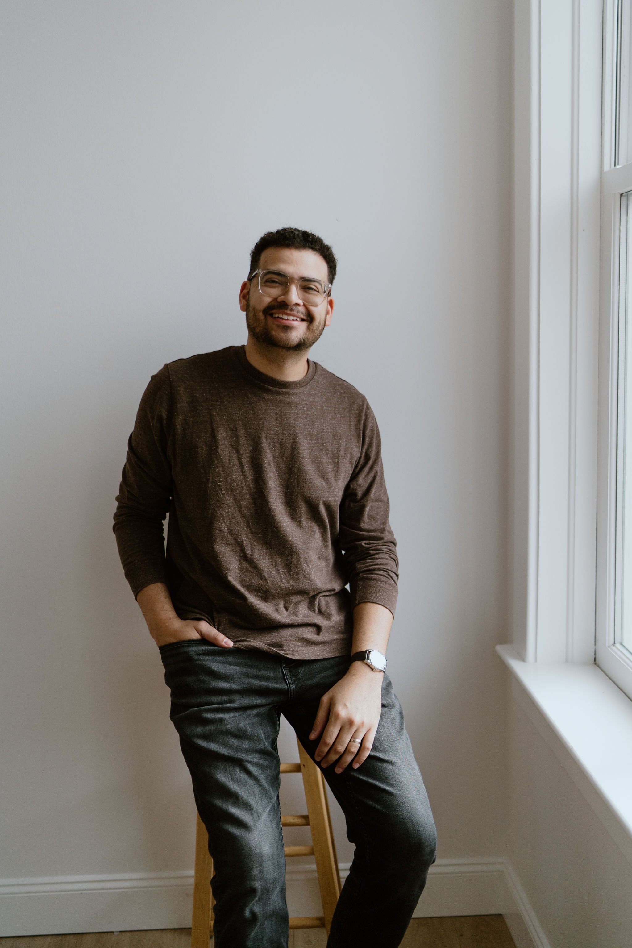 Smiling man with glasses, beard, wearing brown sweater and jeans, sitting on a wooden stool near a large window with white frame sunlight streaming in, against a plain light-colored wall.
