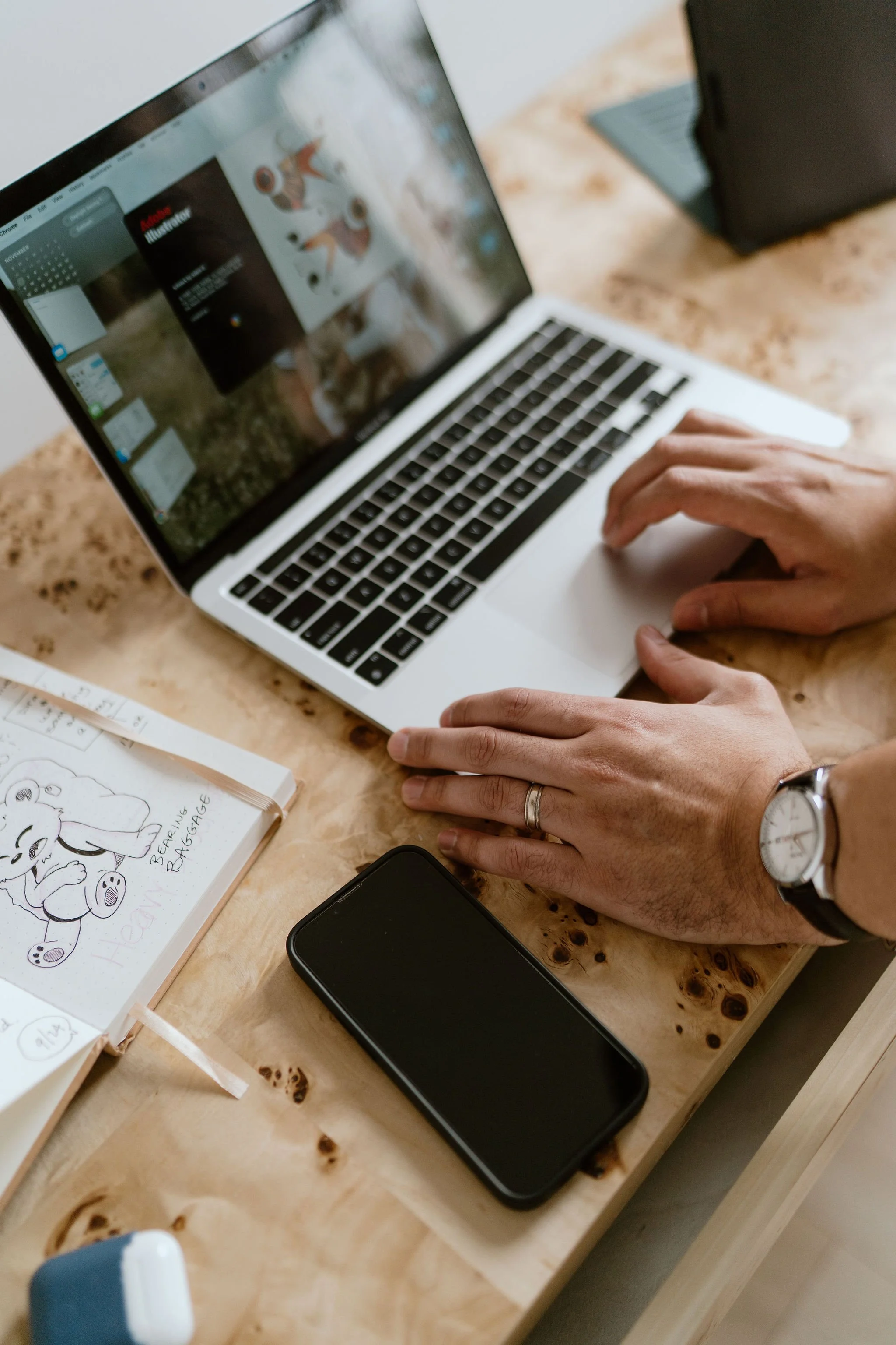 A person working on a MacBook laptop on a wooden desk, with a smartphone and a notebook nearby.