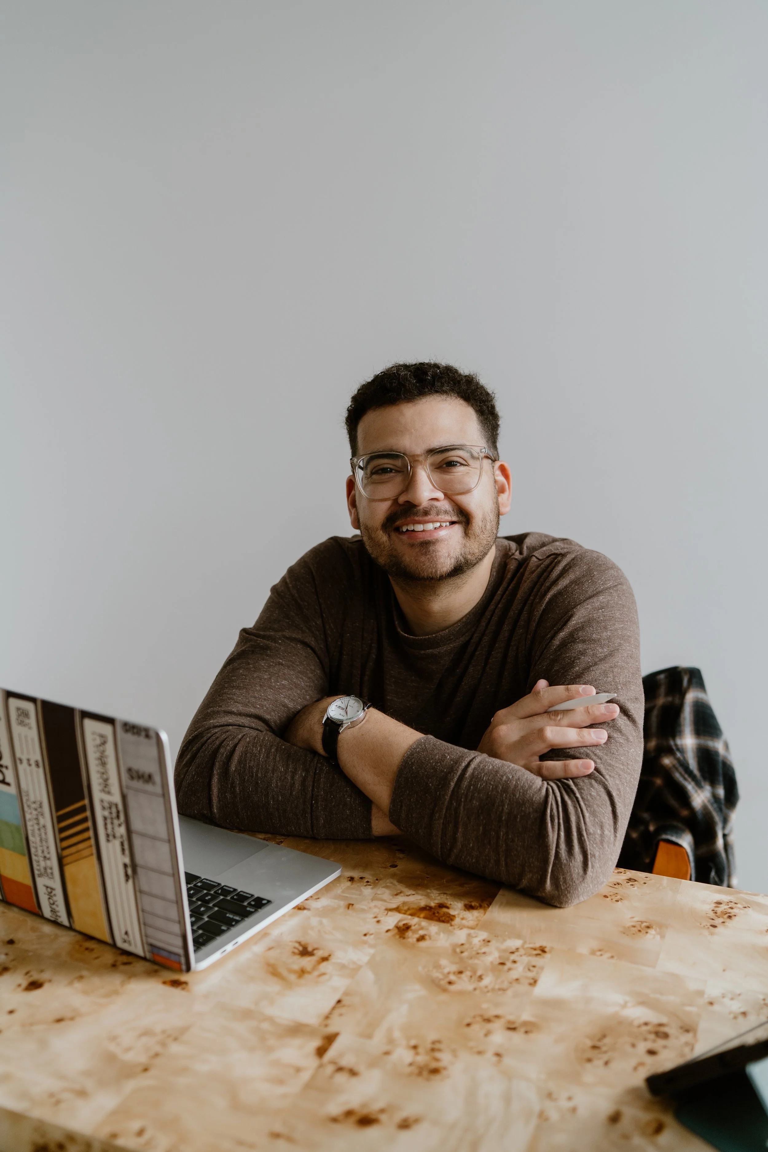 A man with glasses and a watch sitting at a wooden table with a laptop and smiling.