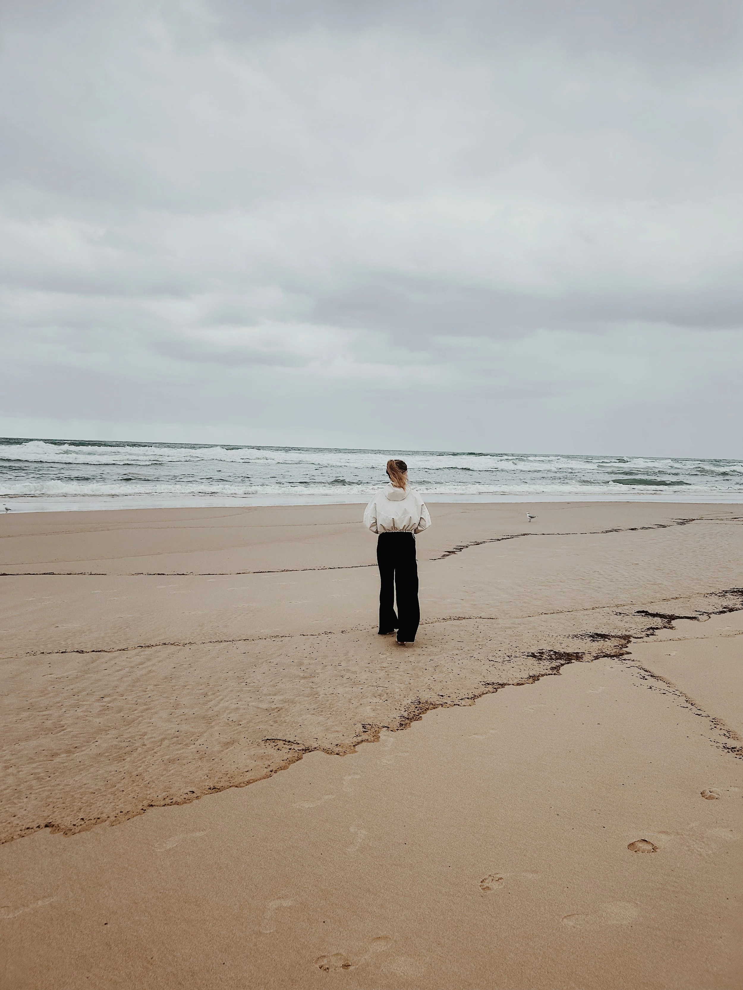 Eine Person steht am Strand, blickt aufs Meer, bei bewölktem Himmel.
