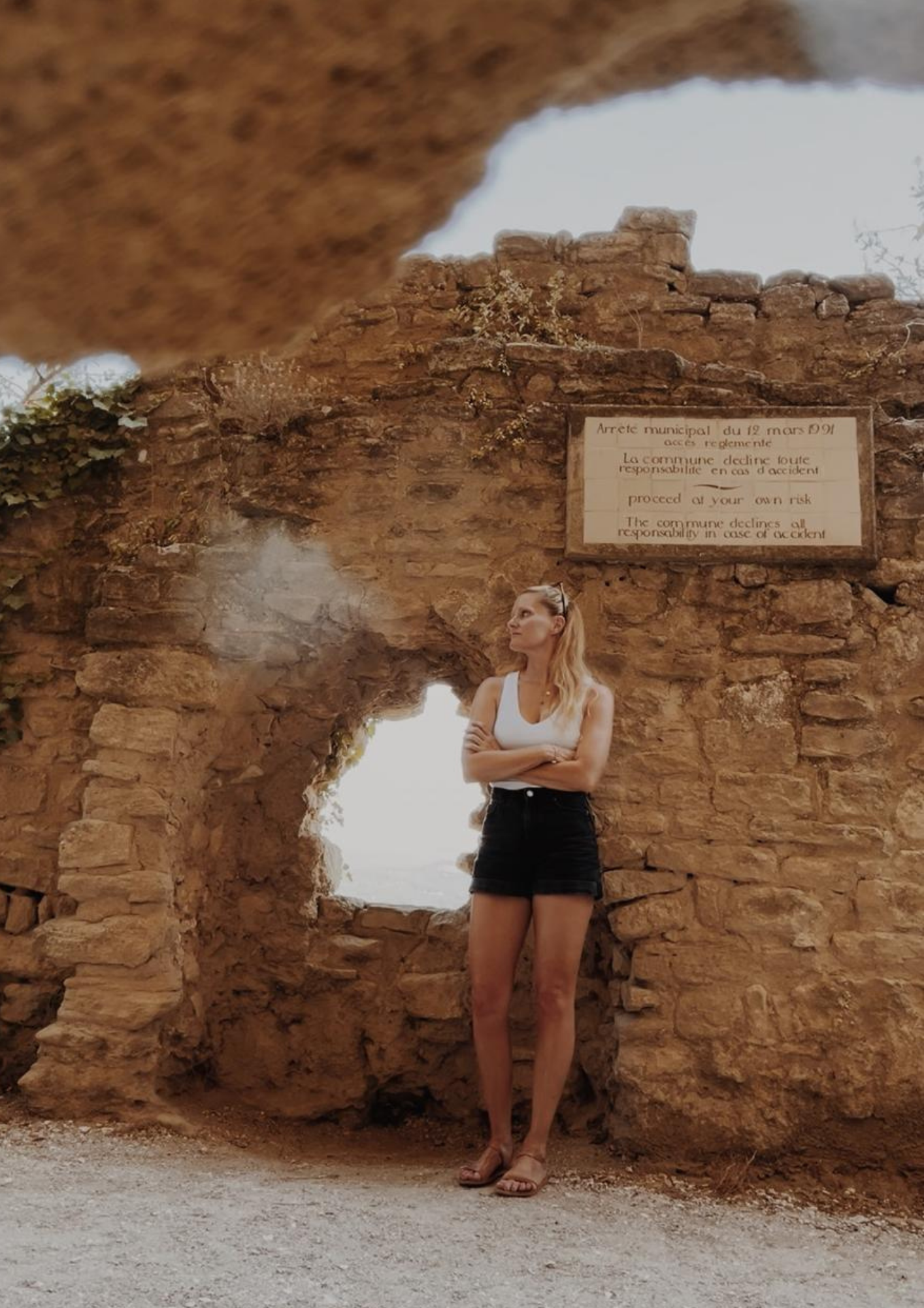 Junge Frau steht vor einer alten Steimauer mit einem Schild, das auf Französisch und Englisch vor einem Unfall warnt, bei Sonnenwetter.