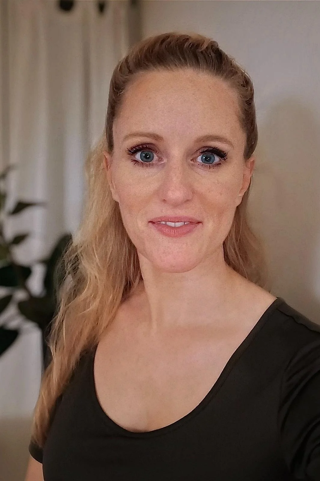 Eine Frau mit blonden, gewellten Haaren, blauen Augen und einem schwarzen Oberteil, die in die Kamera lächelt.