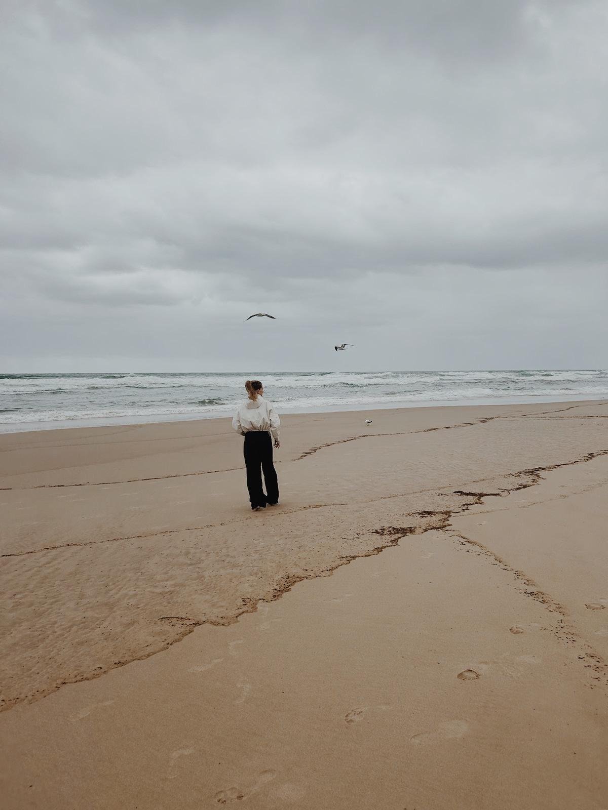 Eine Frau steht alleine am Strand, Blick aufs Meer, über ihr dunkler Himmel mit Wolken, Vögel fliegen über das Wasser, Spuren im Sand im Vordergrund.